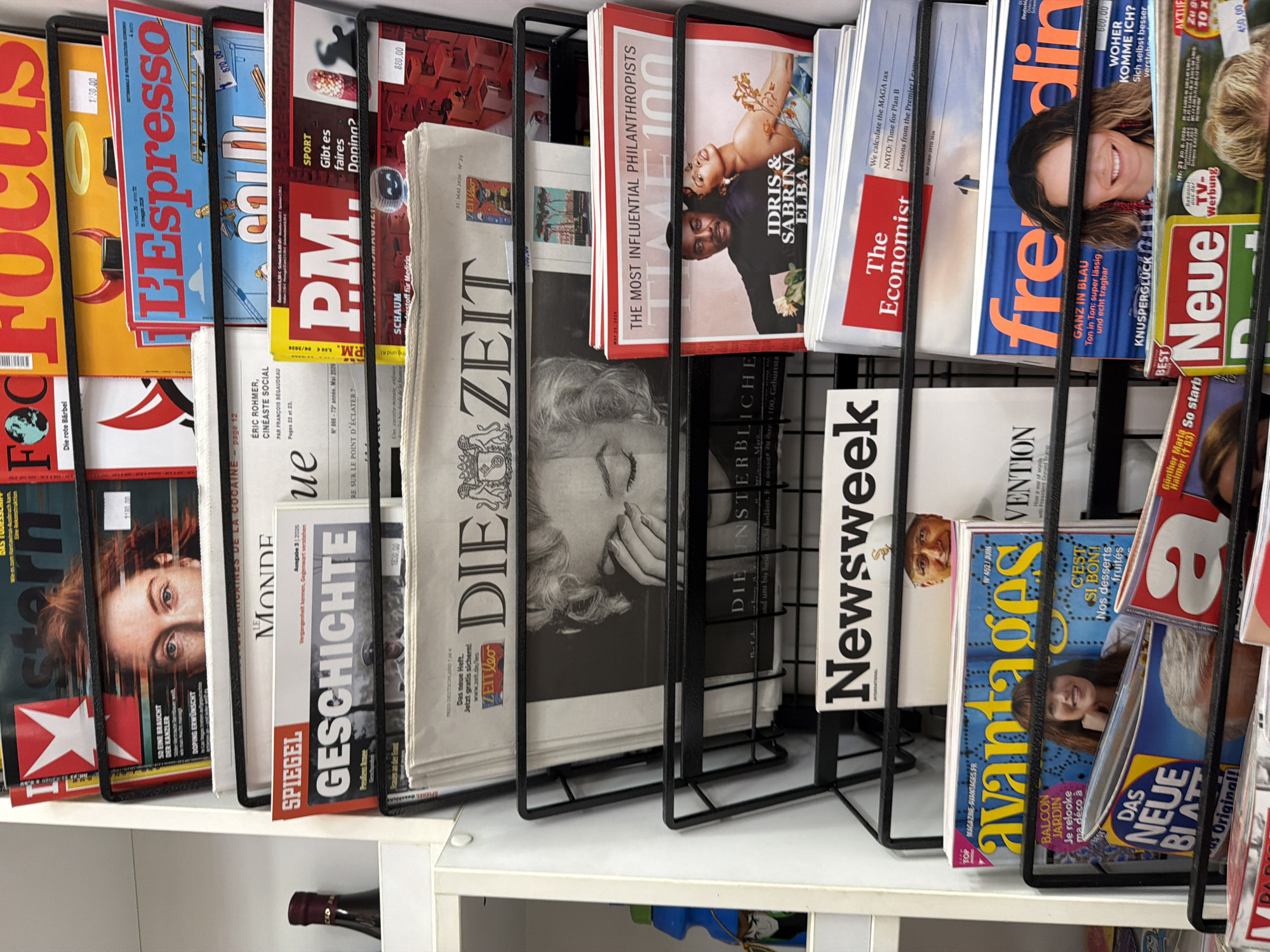 The image shows a magazine rack filled with various publications. The rack is black metal and holds a diverse selection of magazines and newspapers. Visible titles include "Focus", "L'Espresso", "PM", "Spiegel Geschichte", "Die Zeit", "Newsweek", "The Economist", "freedin", "Neue", "TIME", and "avantages". Many of the magazines have people on the cover. The rack is located on a white shelf or cabinet. There is no indication of this image being located in the city of Belgrade, Serbia.