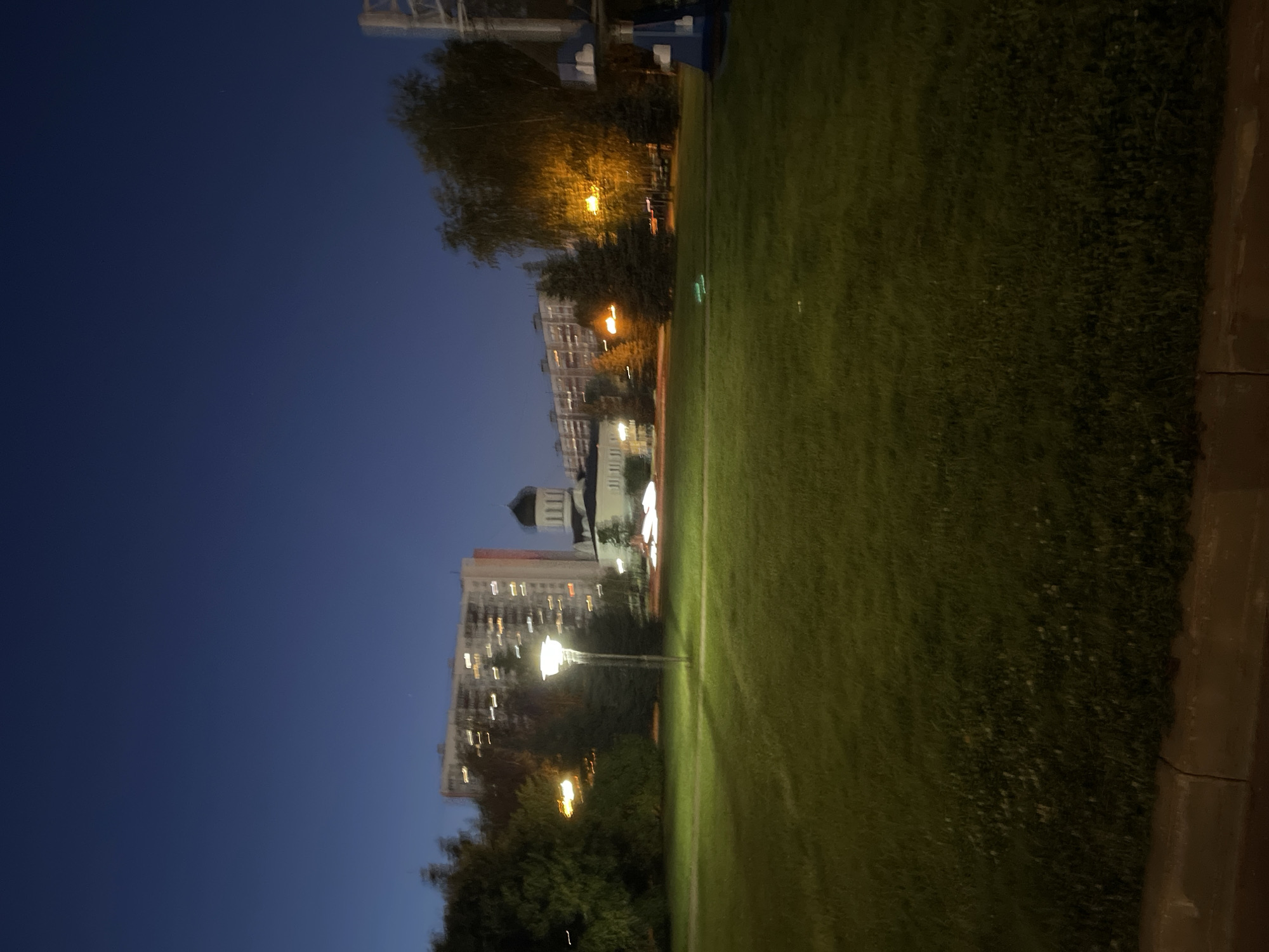 A nighttime, slightly blurred image shows a wide grassy area in the foreground, with a sidewalk or curb along the bottom edge. In the middle ground, a lit-up lamppost stands in the grass, and behind it are several trees and buildings. A domed building, possibly a church, is visible among the trees. Further back, tall apartment buildings with illuminated windows stretch across the horizon under a dark blue sky. To the right, part of a blue structure with cloud-like decorations is visible, possibly a playground element. Streaks of light suggest moving vehicles on a road in the distance. The overall scene depicts an urban park at dusk or night.