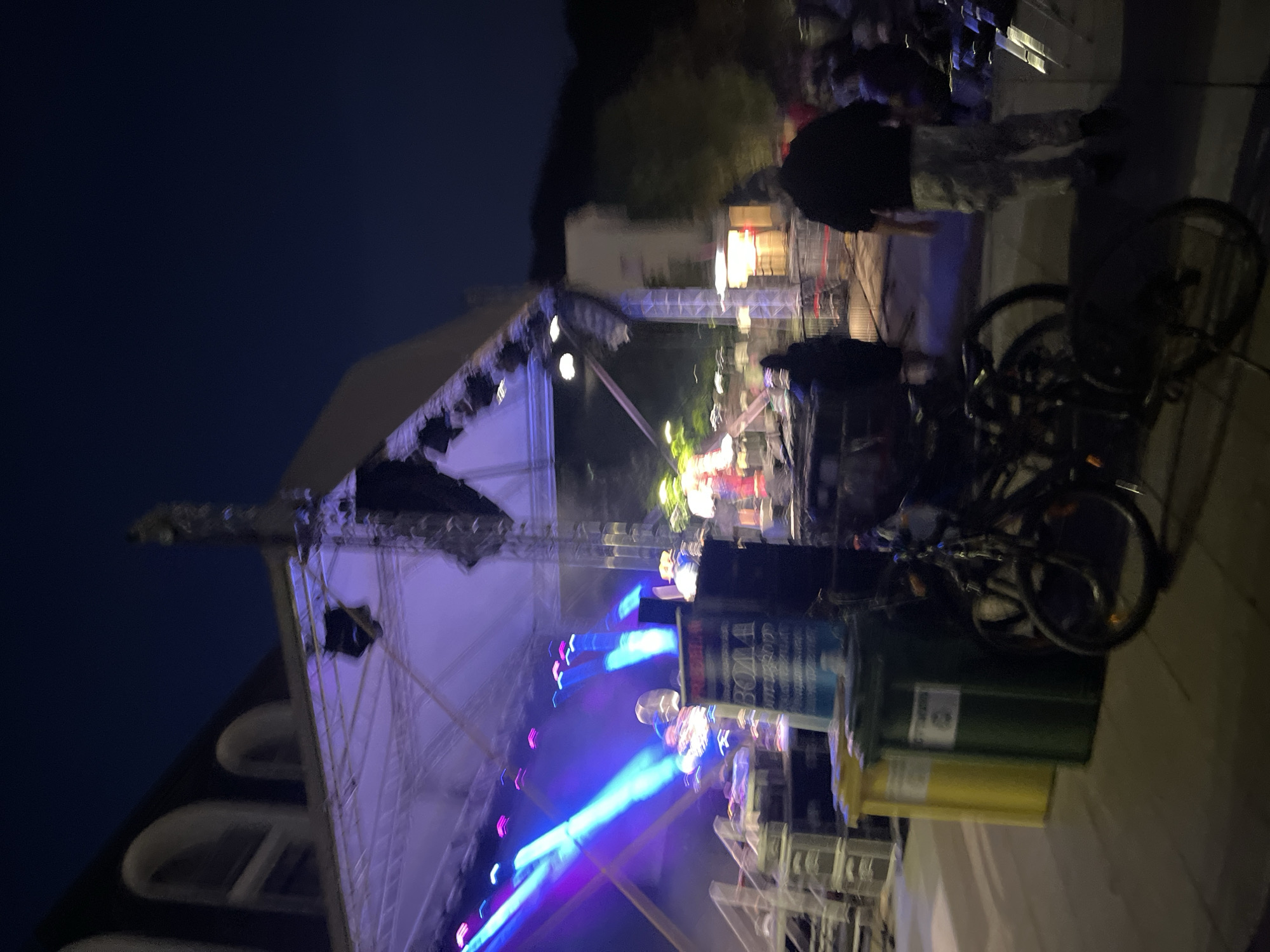 The image depicts a blurry, nighttime outdoor scene, possibly a concert or event. A stage with colorful lights is visible in the background. Several people are present, some walking and others standing. Two bicycles are parked in the foreground, next to two garbage bins, one yellow and one green. The overall atmosphere appears to be lively, with the stage lighting creating a festive mood. The location is unknown.