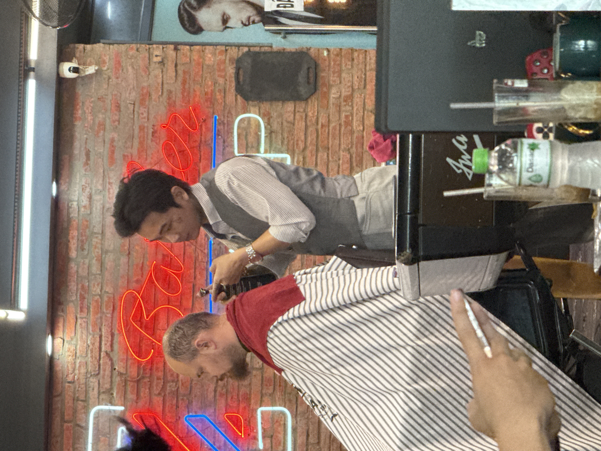 A barber cuts a client's hair. The barber wears a white and gray striped shirt, a gray vest, and a watch on his left wrist. He holds a black instrument in his right hand. The client has a red wrap around his shoulders and is covered in a white and black striped barber cape. A hand holding a white object is also visible. The background features a brick wall with red and blue neon lights and a framed image of a man. A bottle of Dasani water and other various items are visible in front. This scene possibly is located in the city of Đà Nẵng, Vietnam.