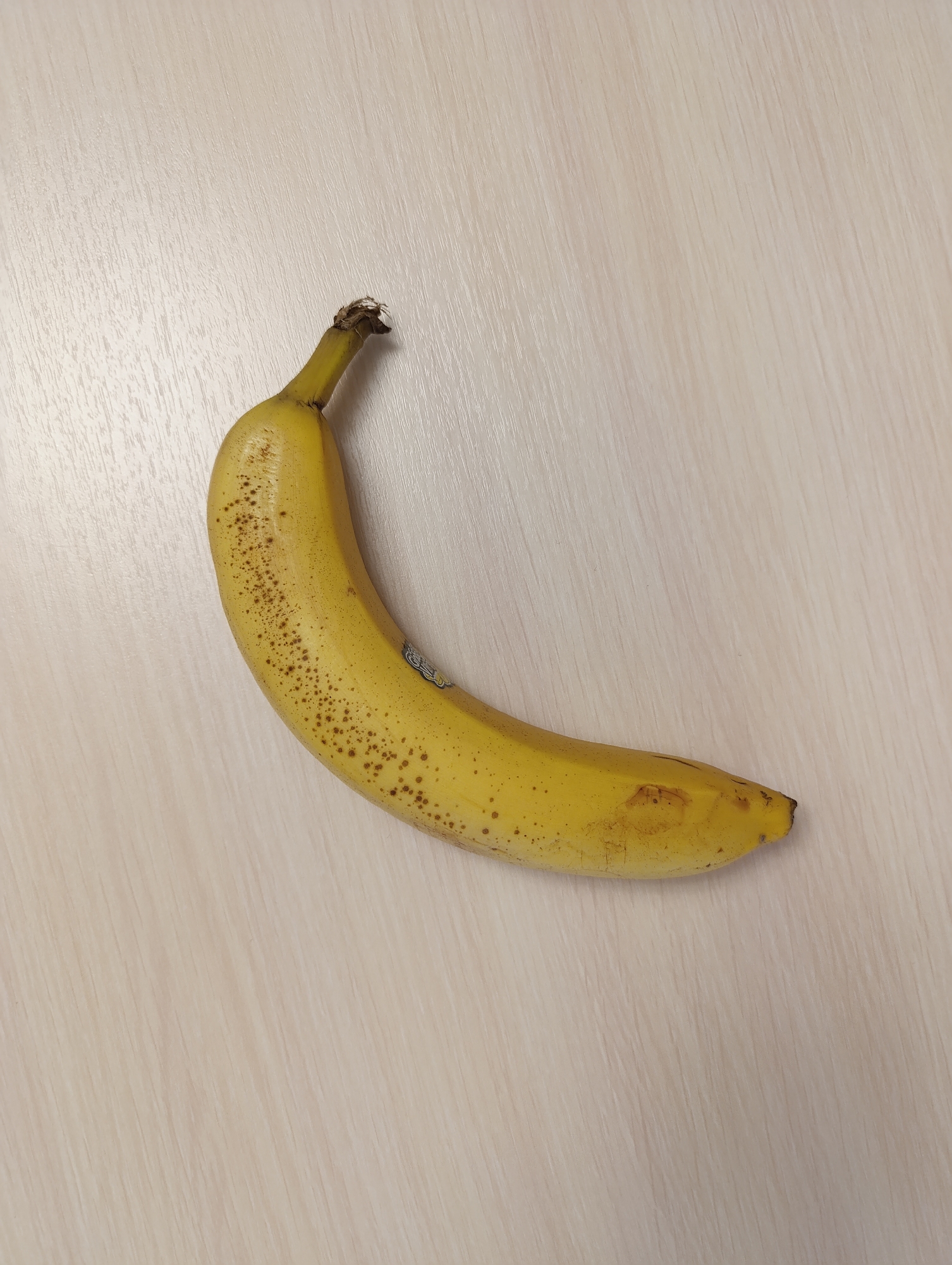 A slightly curved, ripening banana lies on a light beige surface. The banana's peel is mostly yellow with small brown spots scattered across the upper half. The stem end is darker with a dry, fragmented tip. A small sticker is visible on the middle of the banana. The surface appears to be a wood-grain finish. The scene is located in an unknown location within the city of Unknown, Unknown.