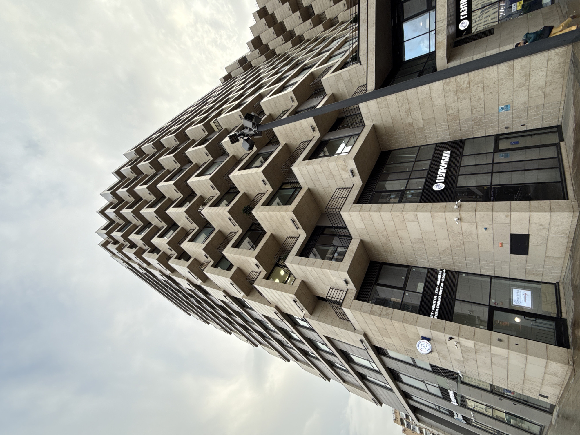 The photograph depicts a tall, modern building in Moscow, Russia. The structure is characterized by a distinctive, stacked-block design with each level featuring projecting balconies or extensions. The building's facade is predominantly clad in a light-colored stone or concrete material, giving it a textured appearance.
Several commercial businesses occupy the lower levels of the building, including a branch of "Gazprombank" (a Russian bank) and "Gestedental", visible through window signage. Other signs indicate medical diagnostic services are available.
The image is taken from a low-angle perspective, emphasizing the height and architectural features of the building against a slightly overcast sky. A surveillance camera is visible attached to the building.