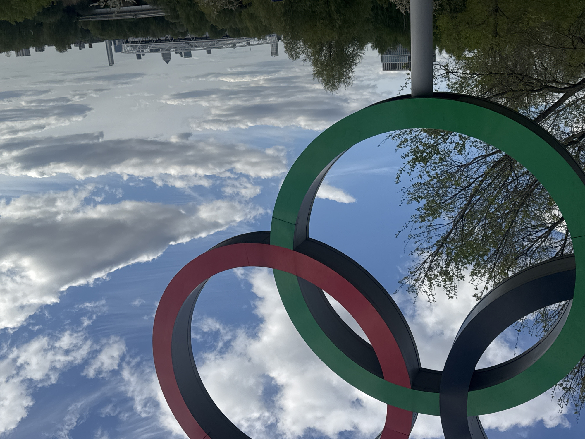 A low-angle, eye-level shot depicts the Olympic rings sculpture in London, United Kingdom, against a backdrop of sky and distant cityscape. The Olympic rings, colored green, red, and black, are the primary focus, with parts of each ring visible. The rings are constructed of metal.
The sky behind the rings is blue, interspersed with white clouds. In the distance, there's a landscape including trees and buildings, suggesting an urban park setting. The image appears to be rotated, giving the impression that the rings are above the viewer's head. The location appears to be near the Olympic Park, London, UK.