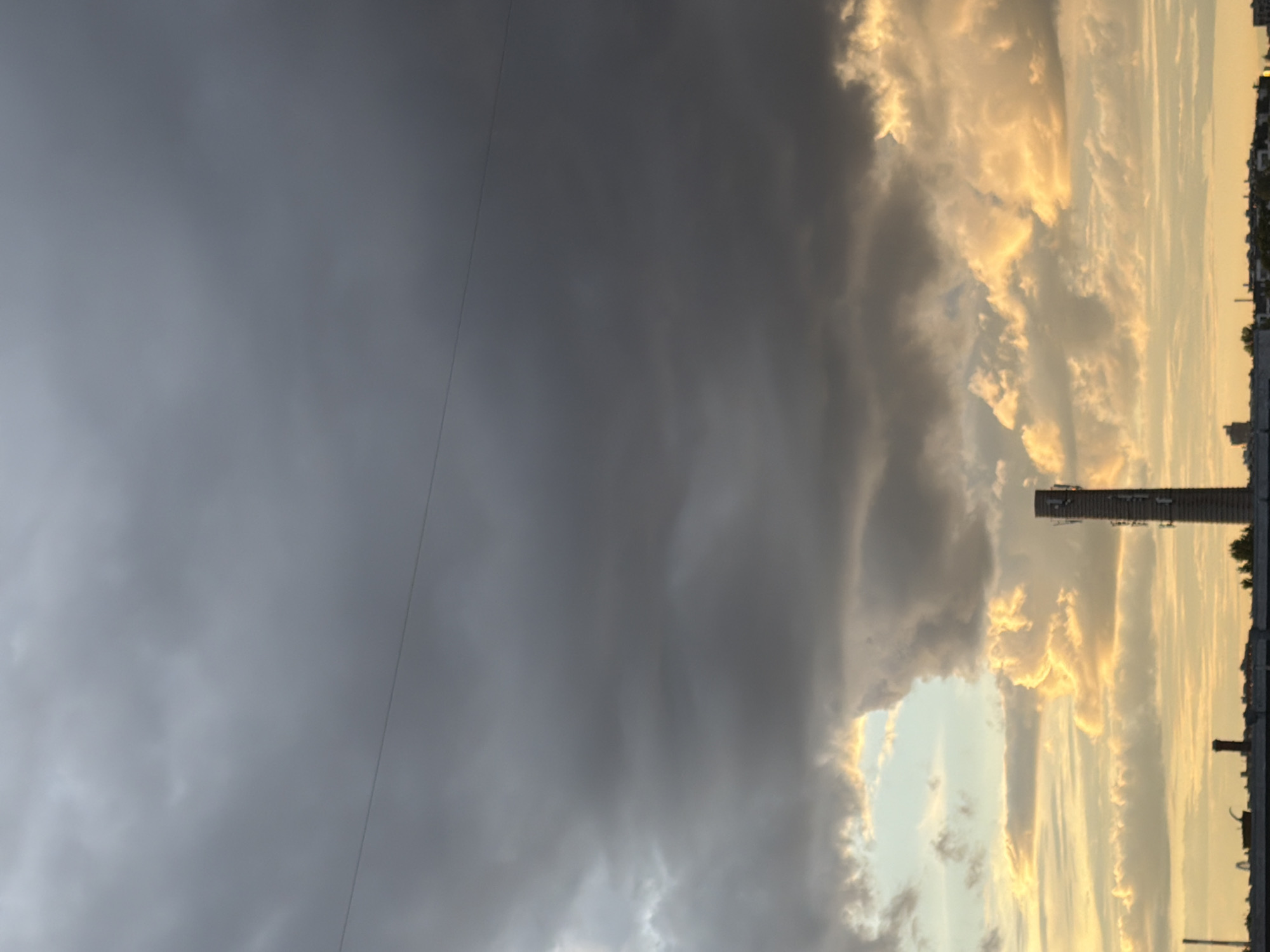 The image captures a dramatic skyscape with a clear division between dark, heavy clouds on the left and brightly lit, wispy clouds on the right. The light illuminating the right side of the sky suggests either sunrise or sunset, casting a warm, golden glow on the clouds. A prominent, tall, rectangular structure, possibly a smokestack or industrial tower, stands against the illuminated sky, topped with what appears to be antennae or broadcasting equipment. Below this structure, a line of buildings with dark rooftops is visible along the horizon, indicating an urban or industrial setting. The contrast between the ominous, dark clouds and the vibrant, sunlit clouds creates a powerful visual effect, hinting at an approaching storm or the clearing of one. A thin, dark line, possibly a wire, runs diagonally across the left side of the frame, adding a subtle detail to the composition. No people are visible in the image. The overall mood is one of nature's power and the dynamic interplay of light and shadow.