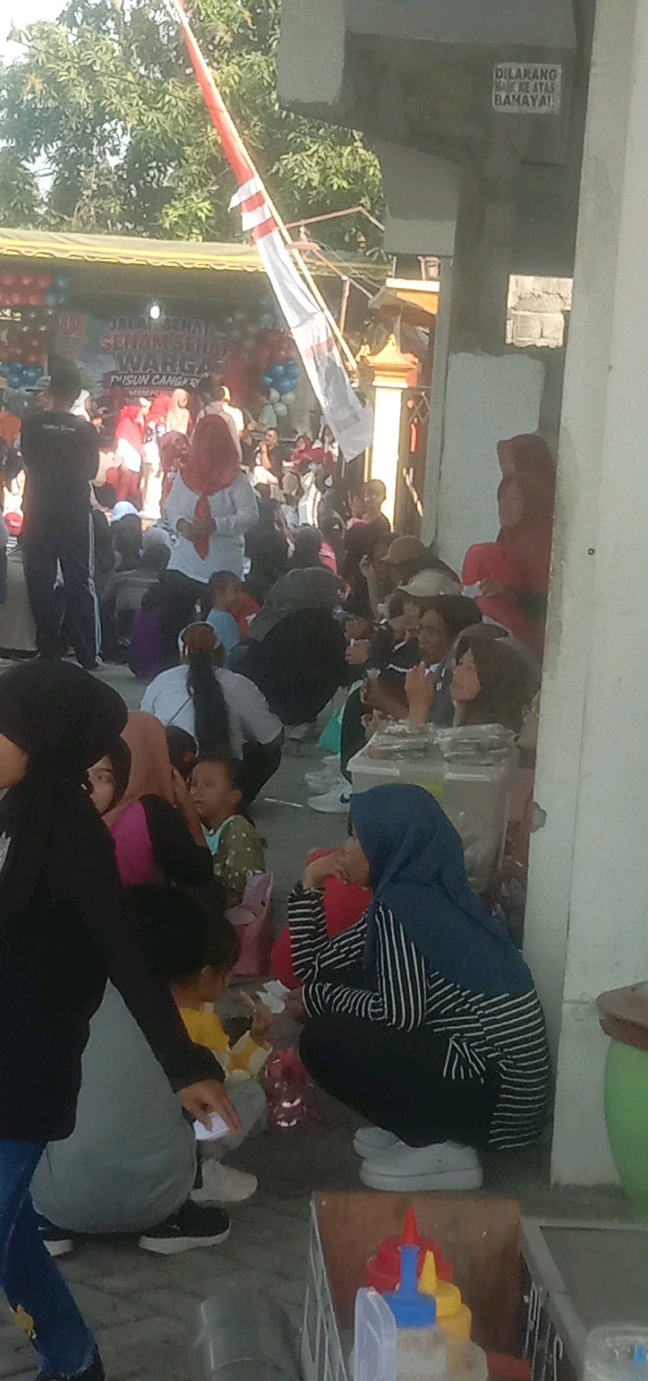The image depicts an outdoor gathering scene in Temuwulan, Indonesia, possibly a community event. In the background, a banner reads "Jalan Sehat Senam Sehat Warga Dusun Cangkring Memper..." suggesting a healthy lifestyle event for the Cangkring Hamlet. A red and white flag is prominently displayed, possibly the Indonesian flag, attached to a pole. Several people are visible, many wearing casual clothing and some wearing headscarves. Some individuals are seated on the ground, while others stand. There appears to be a table with items on it, possibly for sale or distribution, and a cart with condiments. The environment is sunny, with trees visible in the background. One sign on a pillar reads "DILARANG NAIK KE ATAS BAHAYA!" which translates to "DO NOT CLIMB UP, DANGER!". The overall impression is a lively, communal gathering.