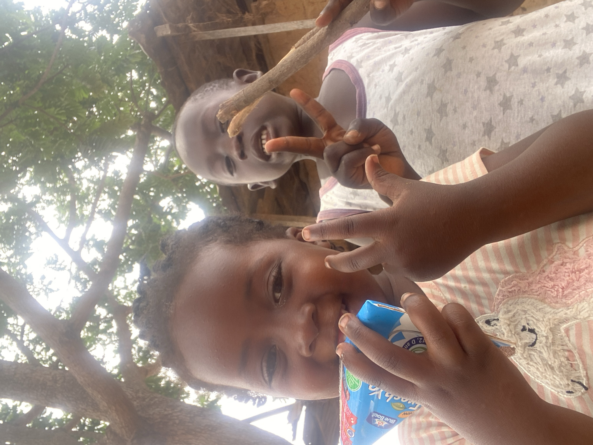The image shows two children in an outdoor environment. The foreground features a child drinking from a blue juice box and making a peace sign. This child is wearing a striped shirt with a unicorn design. Behind them, another child is holding a twig and also making a peace sign. The background includes a tree and what appears to be a simple structure. The location is Kuru Dungus, Nigeria.