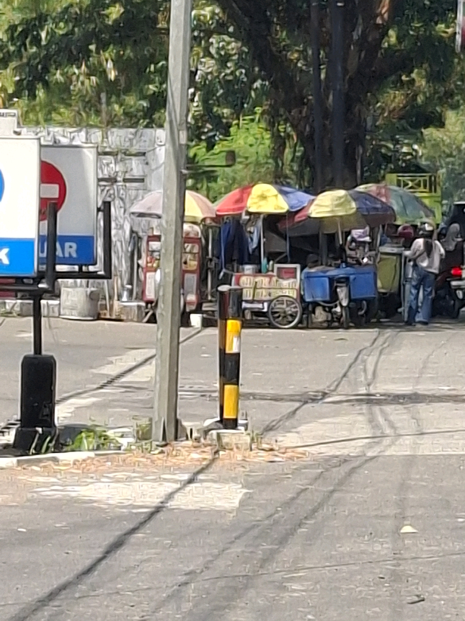 The image depicts a street scene in Cikahuripan, Indonesia, with a focus on a small collection of roadside vendors. In the foreground, a gray asphalt road with tire marks and some scattered debris is visible. A yellow and black striped traffic bollard stands on the right side of the road. To the left, a black metal pole with a black rectangular base supports a sign with a blue background and white lettering that appears to read "JK". Next to this sign is another sign with a white background and a red circle containing a white horizontal bar.
Further back, a row of vendor stalls with colorful umbrellas is set up. The umbrellas are red, yellow, and blue, providing shade for the vendors and their goods. One vendor is operating a small blue cart that appears to be a food or drink cart. A person in blue jeans and a light-colored shirt is standing near this cart, facing away from the camera. Another person in similar attire is seen further to the right, also facing away. Behind the vendors, a large tree with dark bark and green foliage dominates the background, suggesting a well-treed urban or suburban environment. Various structures, possibly small buildings or storage units, are partially visible behind the vendors. The overall impression is a busy but casual street corner with local commerce.