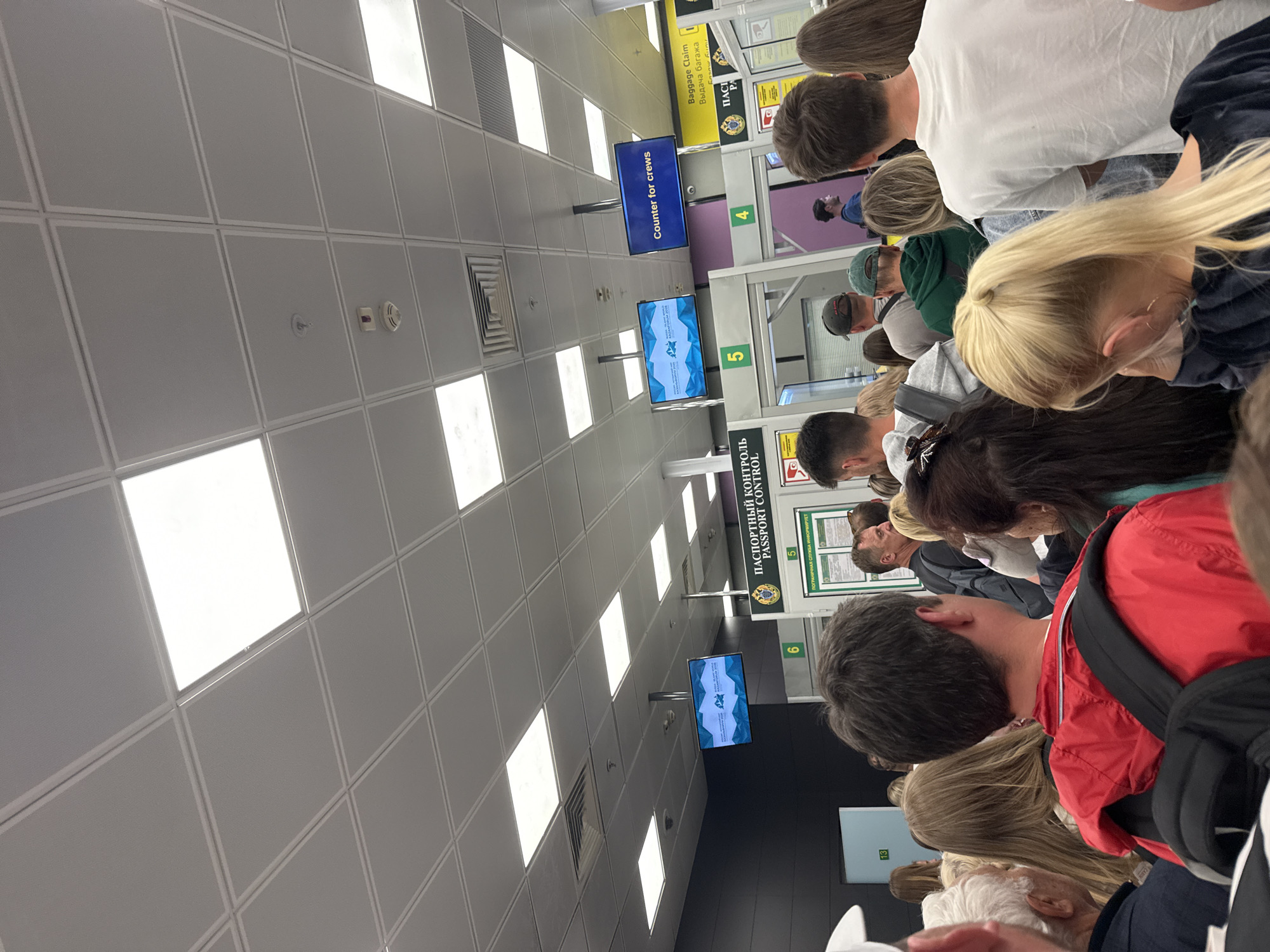 This image shows a crowded line of people in what appears to be an airport, based on the signs visible in the image. Signs in Russian are visible as well as English. "Counter for crews" is one of the signs. The area features a tiled ceiling with fluorescent lights. People are mostly obscured, with only the backs of their heads and some of their clothing visible.