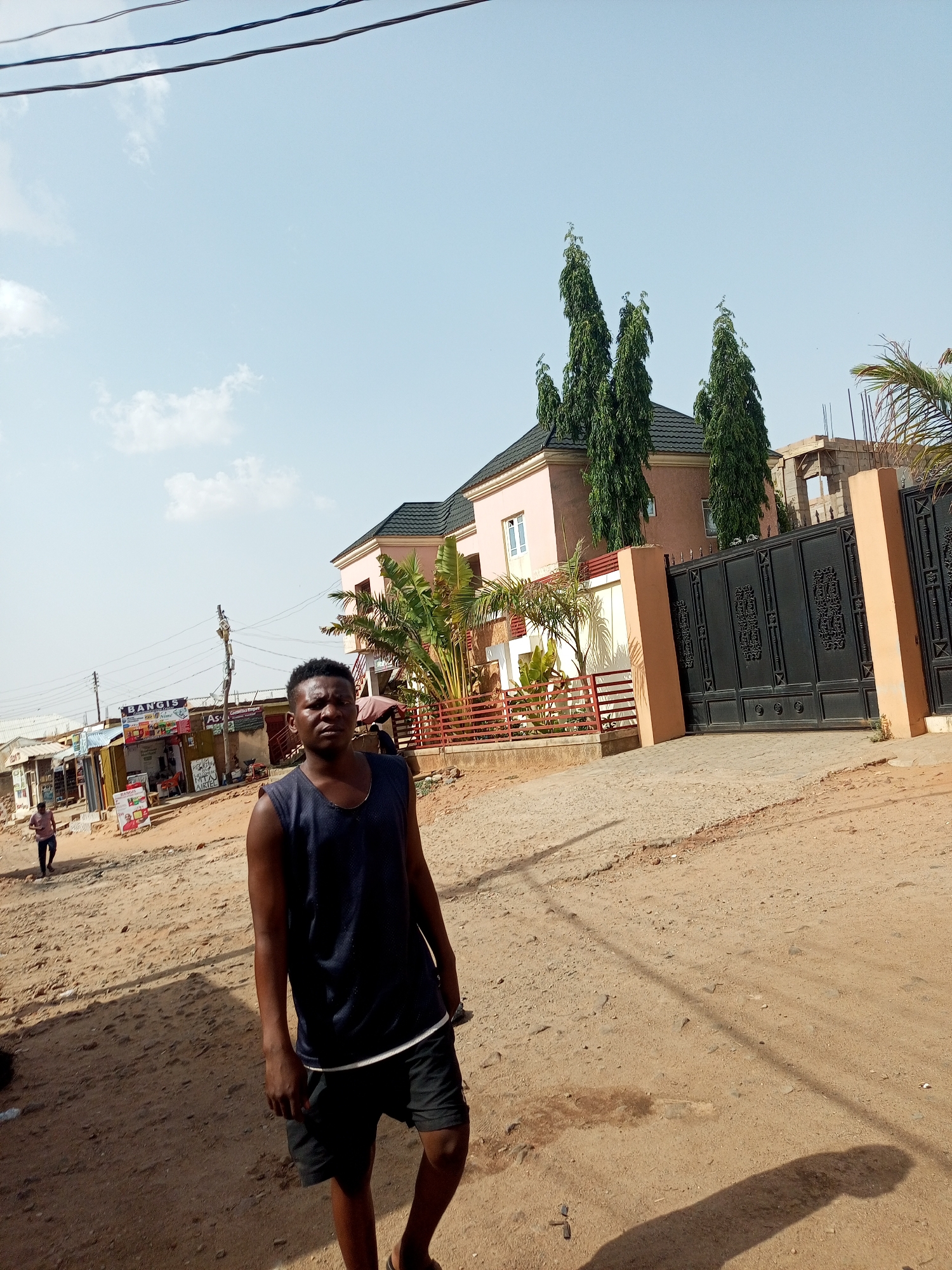 A medium shot of a man standing on a sandy, unpaved road with small stones. The man is wearing a sleeveless dark blue shirt, black shorts, and flip-flops. He appears to be facing the camera, his body slightly turned towards the left. His facial expression is neutral. Behind him is a light peach-colored building with a dark roof, a black gate, and tall, slender trees. In the background, there are various structures, including shops with visible signage. Power lines are visible overhead. The scene is bathed in sunlight, and the sky is light blue with scattered clouds. The location is Tempure, Nigeria.