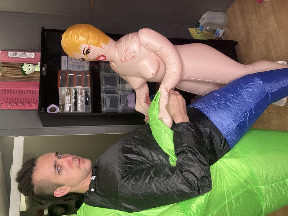 A man lies on his side wearing an inflatable green, black, and blue suit, with his head in the lower left. He looks at the camera. Above him, an inflatable doll with blonde hair and red lips is positioned on a black shelf, appearing to crawl over him. A clear plastic container of supplies is on the shelf behind her. The floor and wall are visible in the background.