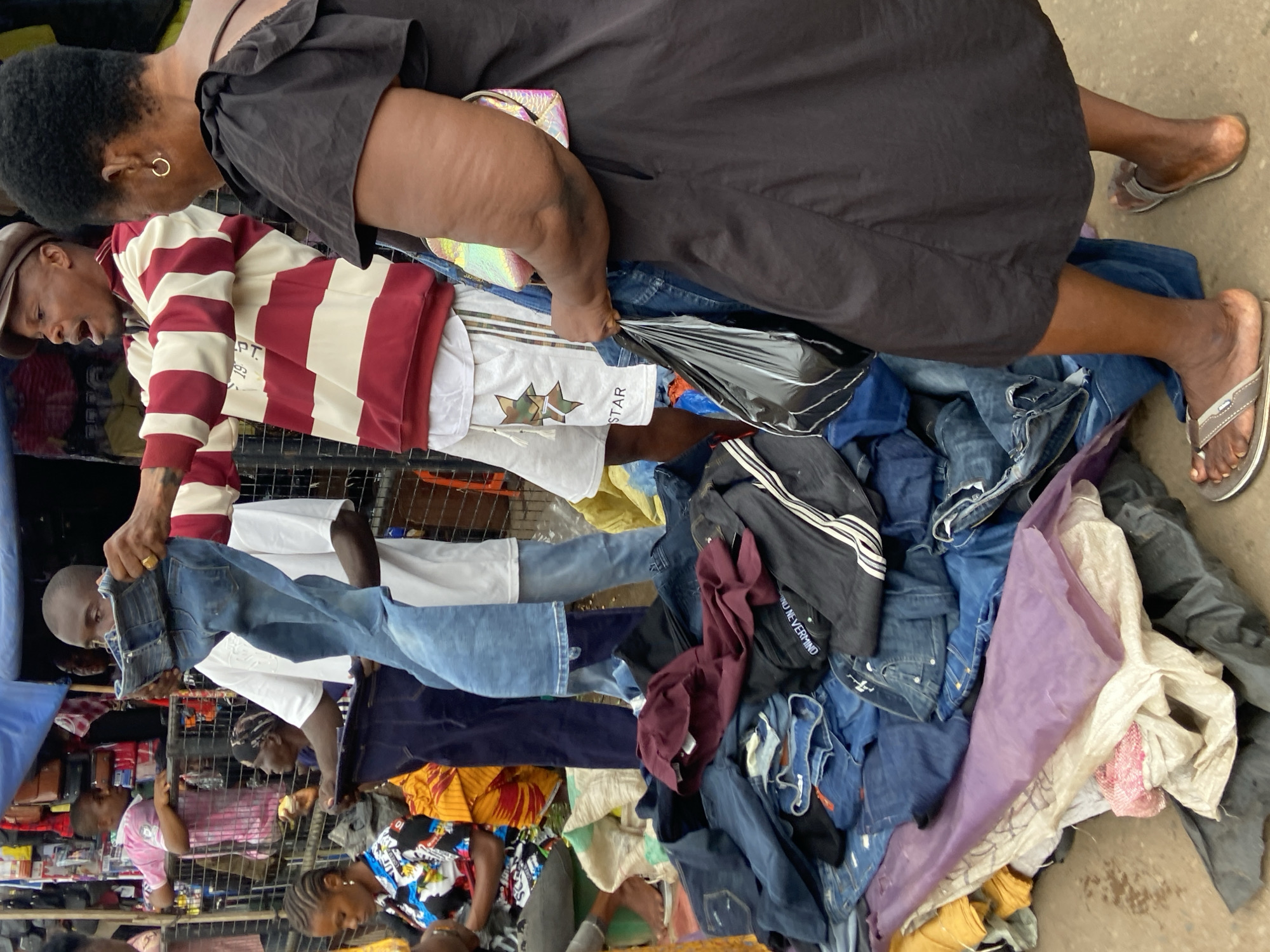 In Nkpor, Nigeria, a vendor stands behind a stall piled high with blue denim jeans. He holds up a pair of jeans, displaying them to a woman in a black dress and a small, iridescent handbag. The vendor wears a red and white striped long-sleeved shirt and white shorts. Behind him, other people are visible in the bustling market, including a young person in a pink shirt and a woman with a headscarf. The ground is covered with more clothing and a few plastic bags. The scene depicts a typical street market in Nkpor, with goods for sale and people interacting.