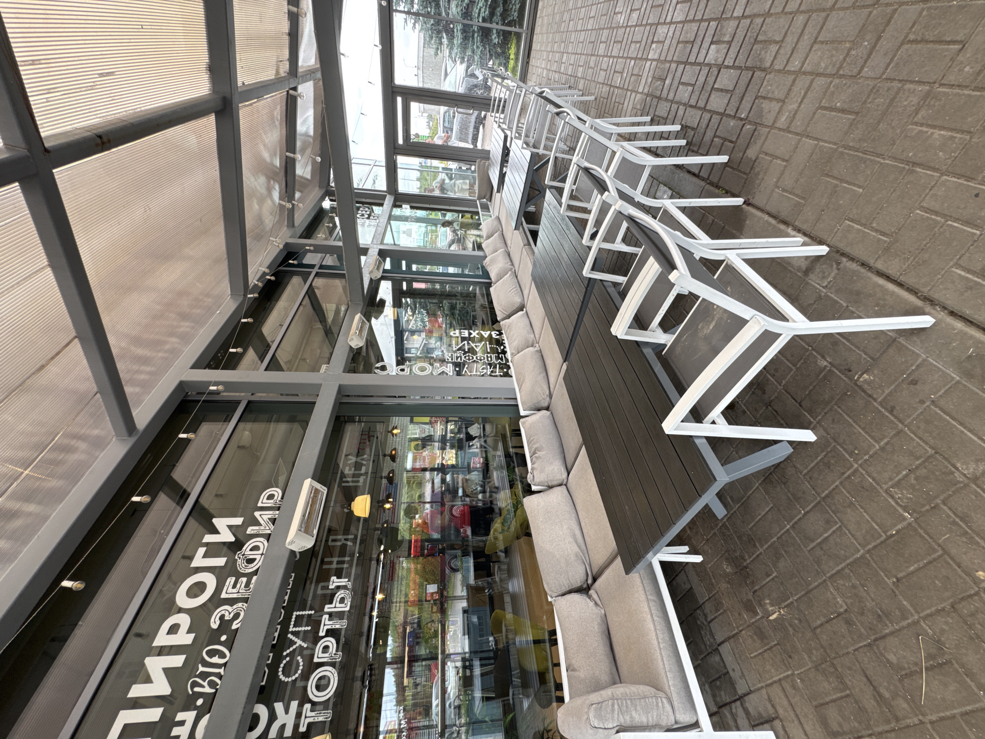This media file shows an outdoor seating area of a cafe or restaurant, likely in Medovoye, Russia, based on the provided context. The scene is viewed from a slightly elevated, wide-angle perspective, with the image rotated so the seating area runs diagonally from the bottom right to the top left.
The main subjects are a series of outdoor tables and sofas, made of dark wood with grey upholstered cushions, and white metal frames. There are also several white metal chairs with grey seats stacked or positioned around the tables. The furniture is arranged on a paved area made of grey rectangular bricks.
Behind the seating area is a glass-enclosed structure, possibly the interior of the establishment. Through the glass, reflections and interior details can be seen, including people, shelves, and lighting fixtures. There is also text visible on the glass, written in white Cyrillic letters. Some legible phrases include "ПИРОГИ" (Pies), "ВІО. ЗЕФИР" (Vio. Marshmallow), "ТОРТЫ" (Cakes), and "СУП" (Soup). Another visible text appears to be "TASTY. MORS".
The time of day appears to be daytime, possibly overcast, as there are no strong shadows and the light is diffused. The weather seems to be mild, as people are not visible wearing heavy clothing. The overall atmosphere is one of an empty, ready-to-be-used outdoor dining space. There are no people actively engaged in activities within the frame. The scene is well-lit, with the grey overcast sky contributing to the ambient light.