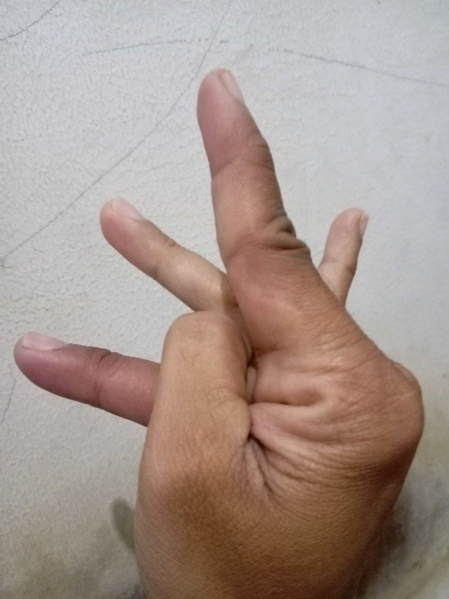 A close-up, high-angle, indoor shot shows a left hand against a textured, light-colored wall. The hand is positioned to the right of the frame, with the fingers extended and bent. Specifically, the index finger and middle finger are extended upwards and slightly to the left, forming a V-shape. The thumb is tucked in, and the ring finger and pinky finger are curled downwards, also tucked in. The skin tone of the hand is medium to tan, with visible wrinkles on the knuckles and palm. The wall behind the hand appears to be a textured surface, possibly concrete or plaster, with subtle linear patterns. The lighting is relatively uniform, casting soft shadows. The scene is devoid of any other identifiable objects or individuals. The provided image does not contain any visual cues that would allow for the determination of its specific location within the city of Batam, Indonesia.