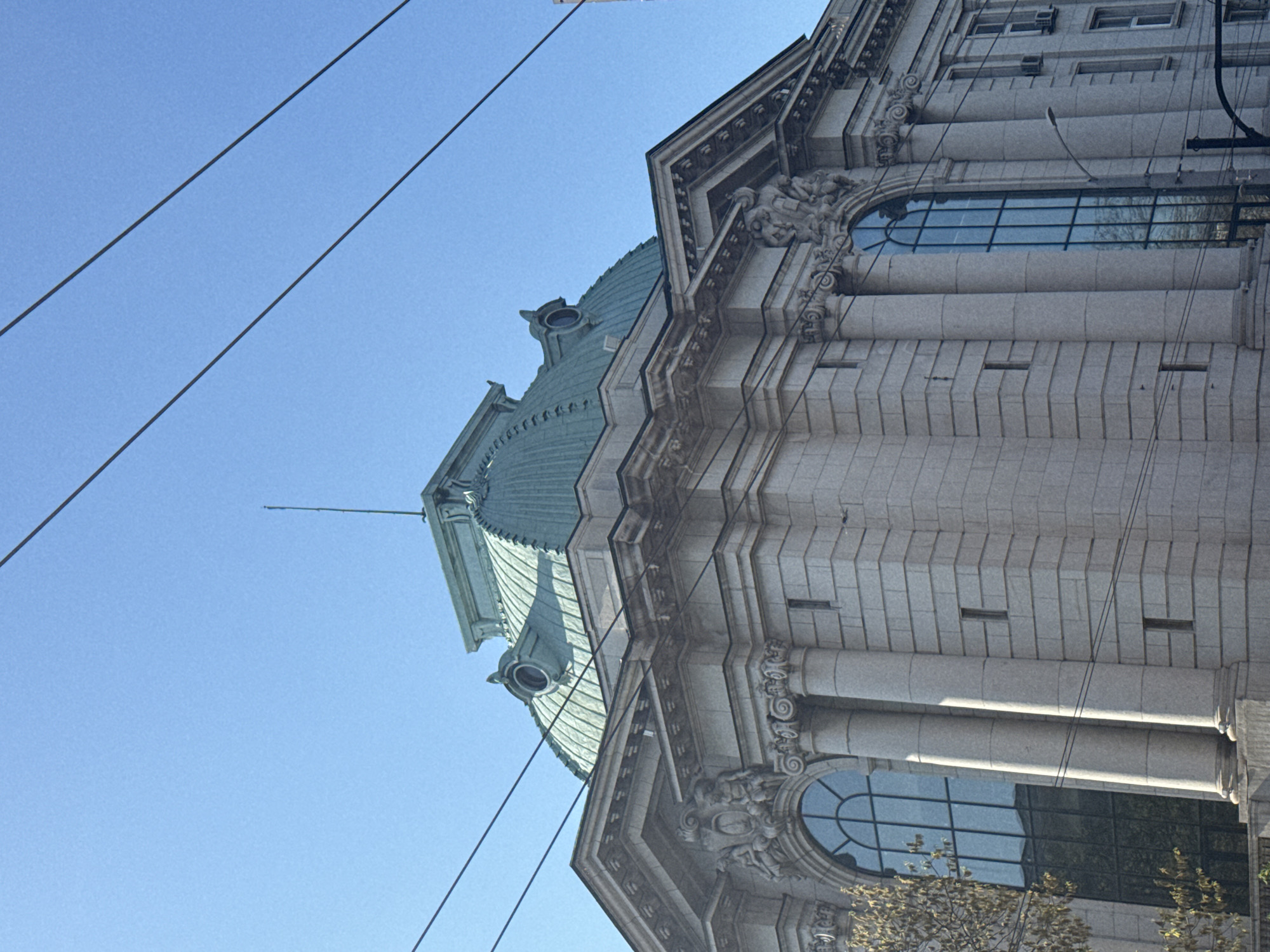 The image shows a portion of the Central Military Club building in Sofia, Bulgaria, focusing on the facade and part of the building's dome. The building is predominantly white with visible architectural details such as columns, decorative carvings, and large windows. The dome has a green hue. Overhead power lines cross the view, obscuring parts of the building and the sky. The sky is clear blue. No people are visible.