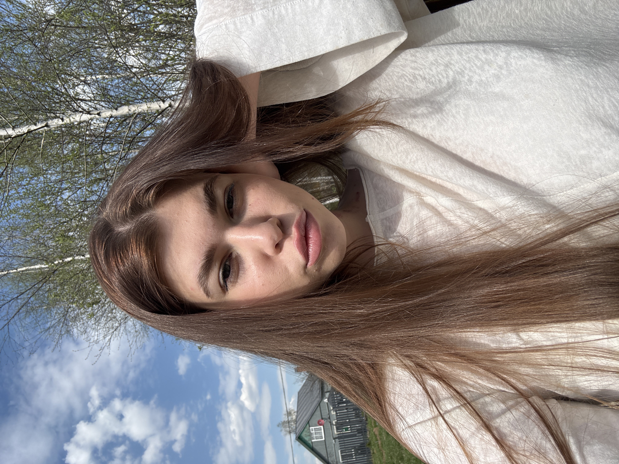A young woman with long brown hair is taking a selfie. She is wearing a white shirt. Behind her is a bright blue sky with white clouds and a tree with thin branches. A small, gray house with a green roof is visible in the lower right corner. The location is likely Шевлягино, Russia based on the prompt.