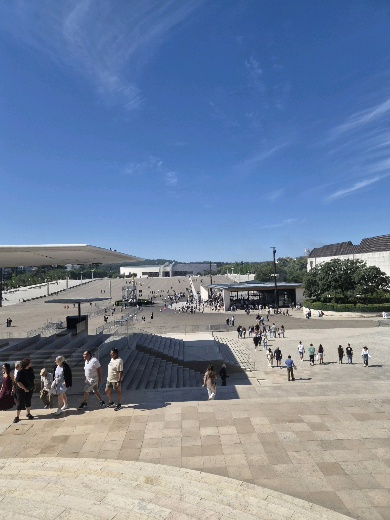 This image shows a wide view of the Sanctuary of Our Lady of Fátima in Fátima, Portugal, on a clear, sunny day. The foreground features large, light-colored stone steps and walkways with a checkered pattern. A group of people are seen walking down the steps, most dressed in casual attire. Beyond the steps, a vast, open plaza stretches out, dotted with many individuals walking or standing.
In the mid-ground, there is a modern, rectangular building with a dark roof and large glass windows, serving as an entrance or exit point for a section of the plaza. To the right, a large, mature tree with dark green foliage stands prominently in front of a long, light-colored building with a dark, tiled roof. Further in the distance, more structures and buildings are visible, extending across the landscape. A large, white, cantilevered canopy extends over a portion of the plaza in the left mid-ground. The sky is a bright, clear blue with scattered, wispy white clouds. The overall scene depicts a public gathering space, likely a religious or tourist site, with a significant number of visitors.
