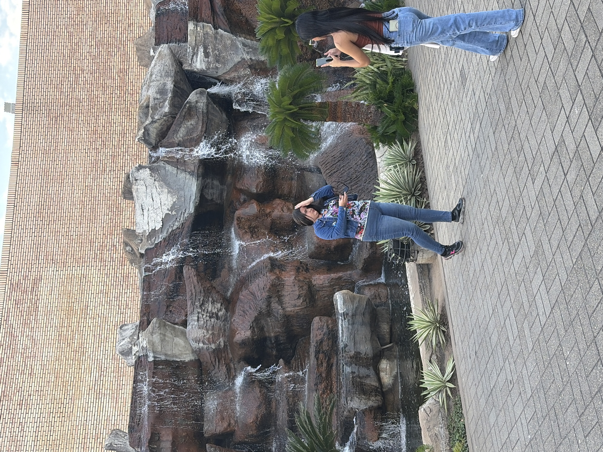 This image captures an outdoor scene featuring an artificial rock waterfall, a paved walkway, and a brick building in what appears to be a public or commercial area in Guayana City, Venezuela.
The central feature is a multi-tiered artificial waterfall constructed from dark, rugged-looking rocks, with water actively flowing and splashing down. Various green plants, possibly cycads or similar palm-like species, are interspersed among the rocks and along the edges of the waterfall, adding to the natural aesthetic.
Two women are present in the scene. One woman, positioned closer to the waterfall on the left, has long dark hair and is wearing a blue denim jacket over a colorful, floral-patterned top, and blue jeans. She is standing casually, looking towards the waterfall, with one hand raised near her head. The second woman, further back on the right, also has long dark hair and is dressed in a rust-colored tank top and light blue flared jeans. She is actively engaged with her smartphone, holding it up with both hands, seemingly taking a photograph or video of the waterfall or the surrounding environment.
The women are standing on a light grey paved walkway, which features a pattern of interlocking pavers. In the background, a large, light brown brick building with a repetitive brickwork pattern dominates the upper left portion of the image. A small, distant tower-like structure is faintly visible above the brick building line. The lighting indicates it is daytime, possibly morning or early afternoon, under fair weather conditions with a partly cloudy sky visible at the top. The overall atmosphere suggests a relaxed, leisurely activity, possibly sightseeing or enjoying the public space. No visible text is discernible in the image.