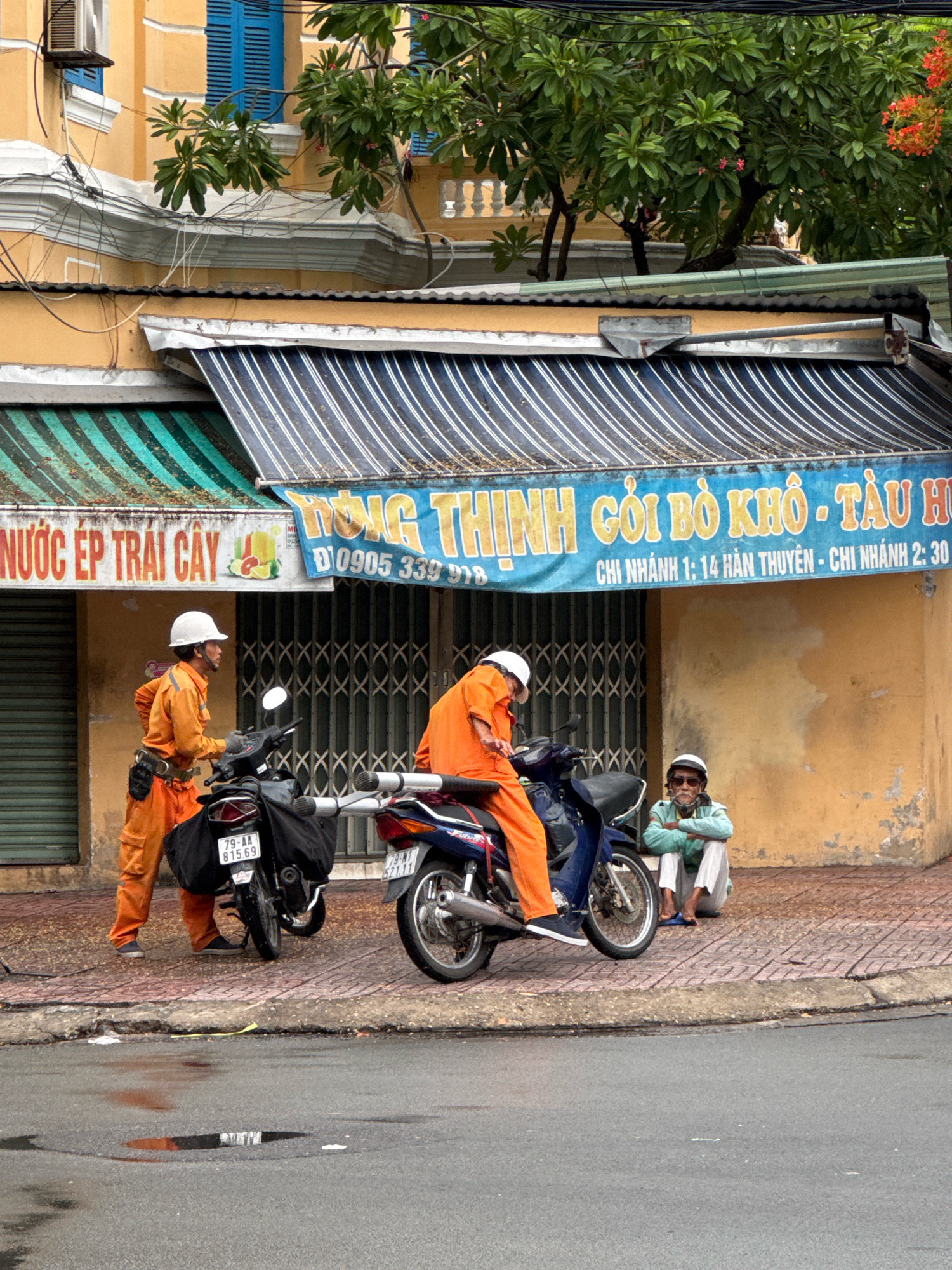 The image shows a street scene in Nha Trang, Vietnam. Two men in orange uniforms and white hard hats are on or near motorcycles. One man is sitting on a blue motorcycle, while the other stands beside a black motorcycle. In the background, an elderly man wearing sunglasses, a light blue jacket, and white pants sits on the ground near a yellow building with a striped awning. The building has a blue shuttered window and a tree with orange flowers. The street is paved with red and grey tiles, and there are puddles on the wet asphalt road. A sign above the awning in Vietnamese reads "Nước Ép Trái Cây" and another sign in yellow lettering reads "HÌNH GÔI BÒ KHO - TÀU HỦ CHI NHÁNH 1: 14 HÀN THUYÊN - CHI NHÁNH 2: 30". License plates on the motorcycles are visible.