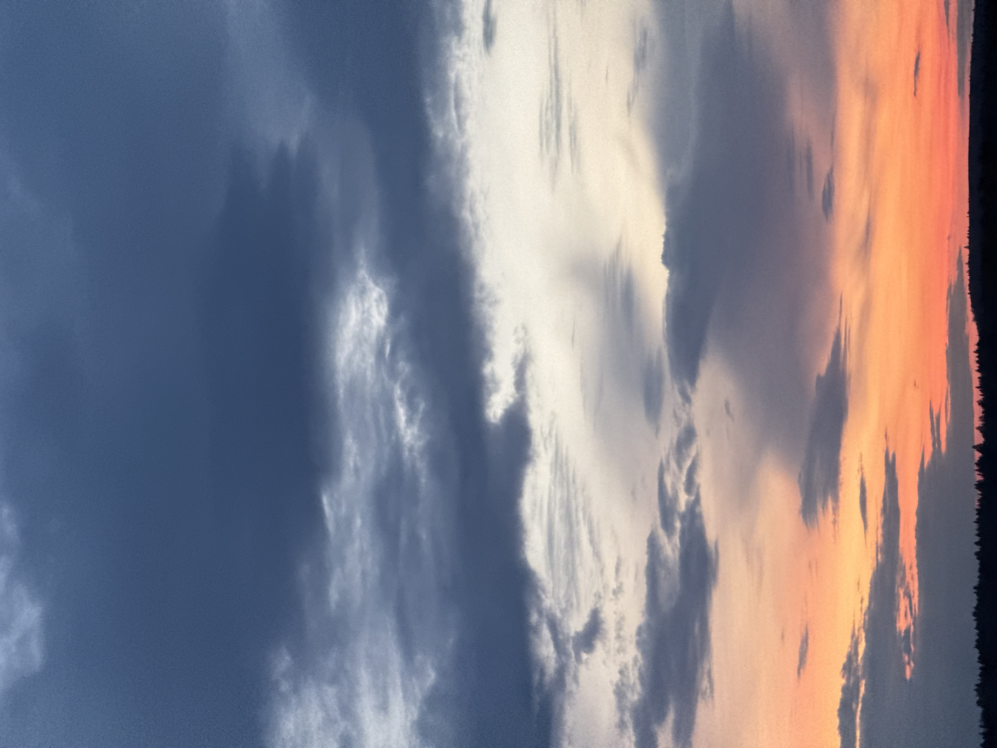 The image shows a sunset sky. The left side of the image depicts dark blue, cloud-filled skies. Moving to the right, the color transitions to lighter gray and white clouds, with a band of orange and pink hues to the far right. A dark silhouette of trees is visible along the top right edge. The image is taken outdoors. The location cannot be determined from the image.