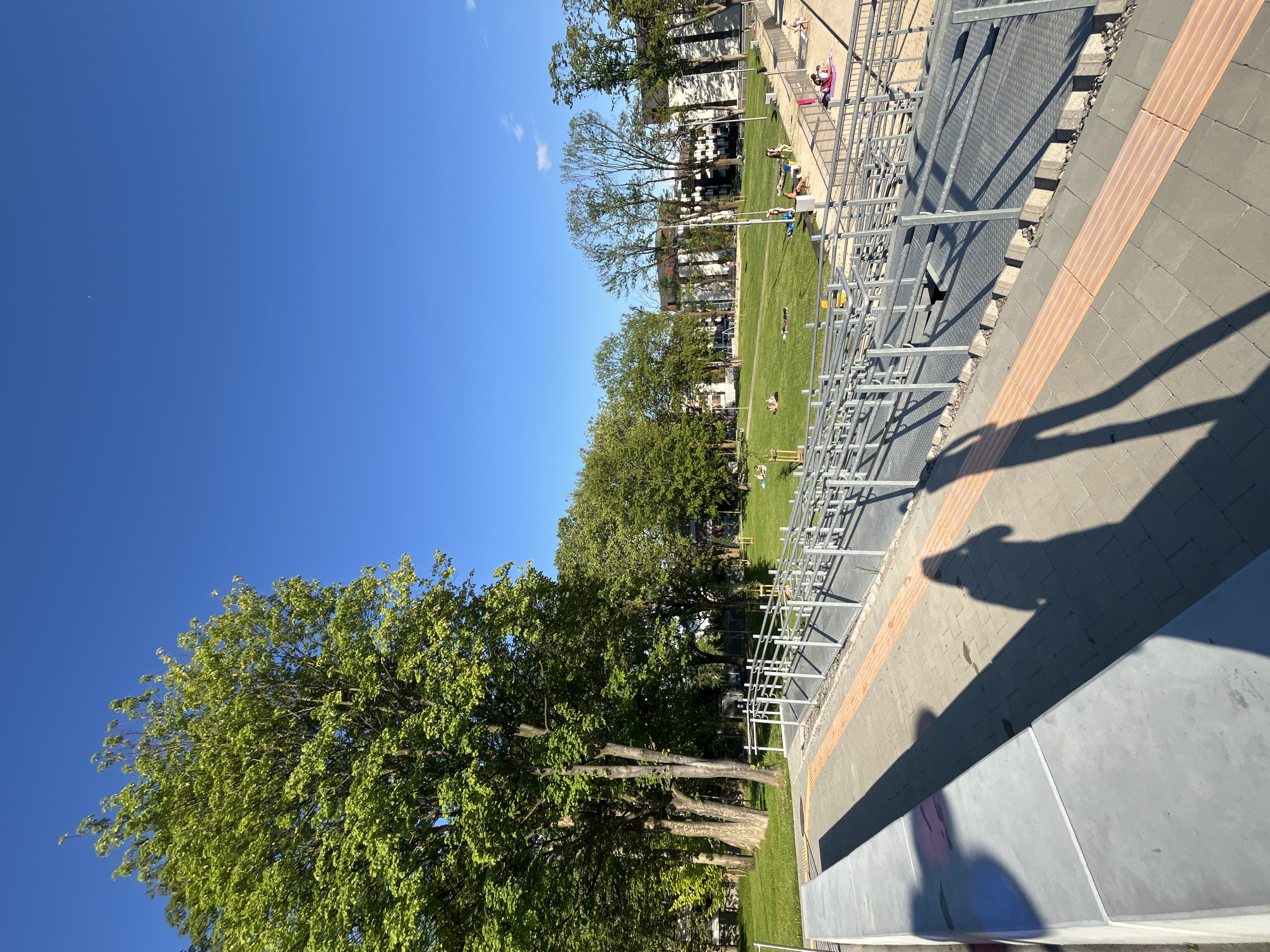 The image depicts an outdoor scene on a clear, sunny day in a public park or green space within the city of Unknown, Unknown. The perspective is from an elevated paved area looking down into the park.
In the immediate foreground, a grey concrete ledge or bench runs along the left edge. The ground is paved with grey rectangular pavers, featuring two parallel strips of orange tactile paving, likely for the visually impaired, running diagonally from left to right across the foreground. Prominent long, dark shadows of two individuals, cast by the sun, stretch across this paved area; one shadow is distinctly human-shaped, facing away from the viewer, while the other is a longer, less defined shadow from a nearby object or person.
To the right of the paved area, a multi-sectioned metal ramp structure with numerous silver railings descends into the park. The ramp's surface is made of a textured grey metal grid.
The midground features a large, expansive green lawn with several mature trees displaying vibrant green foliage, indicating late spring or summer. Scattered across this lawn are approximately 15-20 individuals engaged in leisure activities. Some people are sitting or lying on the grass, seemingly relaxing or sunbathing. To the right of the ramp, a sandy area or beach section is visible, with a group of people gathered, some on towels or mats.
In the background, behind the trees and across the park, several multi-story residential or commercial buildings with light-colored facades are visible. Overhead, the sky is a bright, cloudless blue. A tiny, faint crescent moon is discernible in the upper central portion of the sky. The lighting suggests either early morning or late afternoon, given the length and angle of the shadows.