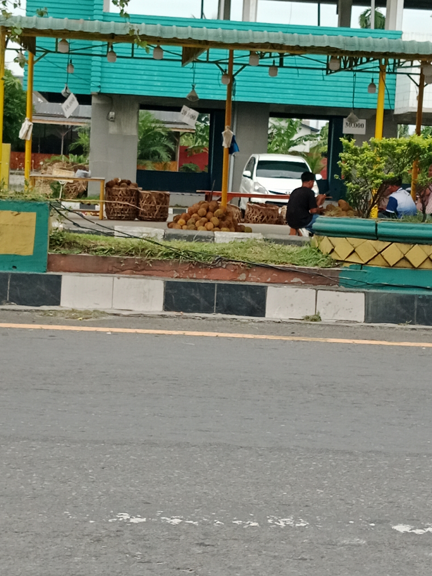 In the city of Binjai, Indonesia, a roadside stall displays durians for sale under a teal and yellow awning. The stall features several woven baskets filled with the spiky fruit, and a large pile of durians is visible on the ground. Two men are present, one wearing a black t-shirt and jeans, crouched near the fruit, and another in a blue and white shirt, sitting down. A white car is parked behind the stall. The area is lined with a decorative concrete border featuring black, white, and red patterns, with green grass and plants. The scene takes place alongside a paved road, with a yellow line marking the edge. Signs with prices like "10.000" and "30.000" are hung from the awning.