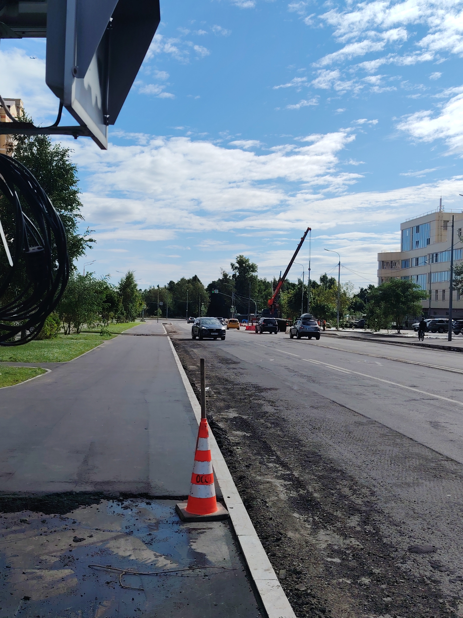 An outdoor, medium-shot image captures a street scene with ongoing road construction in Zelenograd, Russia. The foreground features a paved walkway next to a road undergoing repair, with loose asphalt and a traffic cone with orange and white stripes visible. To the right, the main road displays a mix of paved and rough surfaces, with traffic moving in the distance. Several cars, including a dark SUV and a yellow sedan, are on the road. A large red crane is positioned on the left side of the road further down, suggesting construction work. Green trees line the sides of the road and walkway. In the background, a modern multi-story building with glass windows stands tall. The sky is blue with scattered white clouds, and a small airplane is visible in the upper left portion of the frame. A person on a bicycle is also present on the right side of the road. A black, coiled cable hangs from an unseen structure in the upper left corner.