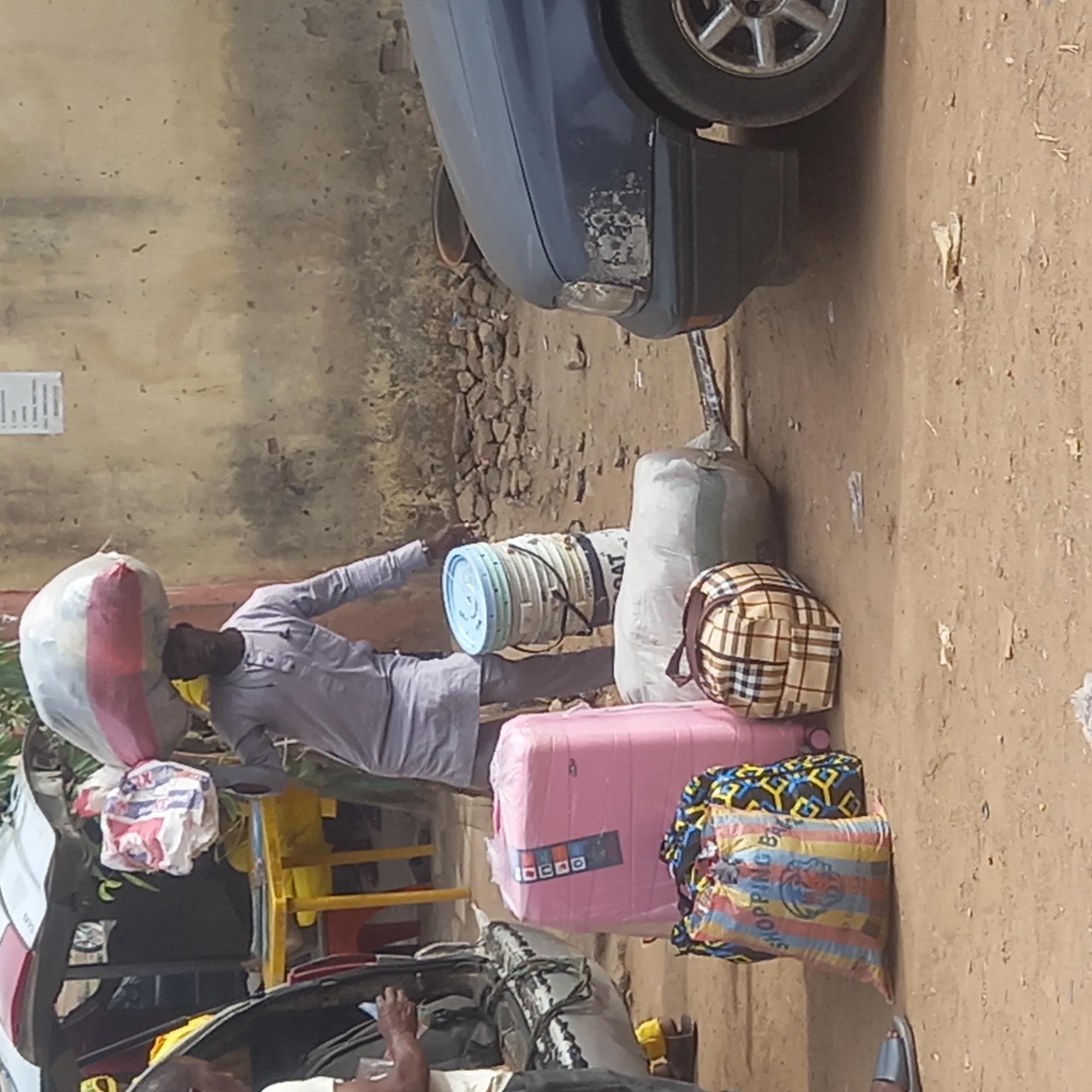 This image captures a busy, informal scene on what appears to be a dirt street or roadside in Sardauna, Nigeria, likely during the daytime. The setting suggests a travel or transport hub where people and goods are being prepared for departure.
In the center, a man, dressed in a light-colored long-sleeved shirt and trousers, stands, skillfully balancing a large, white bag with a red stripe on his head. He extends his left arm, possibly to steady himself or to handle a stack of light blue and white buckets, some bearing the text "CAT," which are tied together and rest on the ground beside him.
Surrounding the man and extending into the foreground and background is a collection of diverse luggage and goods. To the man's right, a large, light-colored sack stands upright. Further right, on the ground, are multiple pieces of luggage: a plaid (brown, white, and blue checkered) backpack, a pink suitcase wrapped in plastic with a "Iocoo" or "Ioeco" label, another bag with yellow, blue, and black geometric patterns, and a colorful striped bag (yellow, red, blue, green) prominently displaying the word "SHOPPING" and a lion head logo.
On the left side of the frame, part of a white and red vehicle, possibly a bus or large taxi, is visible, loaded with more bags and items. In the bottom left corner, a person's hand is seen inside what appears to be another vehicle.
In the background, a weathered, light brown wall with peeling paint provides a rustic backdrop. Below the wall, rough, rocky terrain is visible, leading to the dirt ground where the activity is concentrated. A dark blue car with some visible wear and tear on its bumper is parked further back on the right, with its wheel prominently displayed. A small white paper with illegible text is affixed to the wall. The overall impression is one of people preparing for a journey, amidst a collection of personal belongings and cargo.