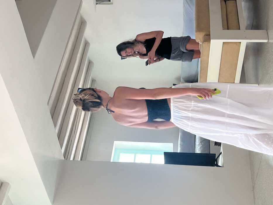 The scene is an interior room within a residence in Tourlos, Greece, characterized by white walls, a light-colored beamed ceiling, and a bright, minimalist aesthetic. Two females are present.
In the immediate foreground, a female is depicted from behind, facing away from the viewer with a slight turn to her right. She has blonde or light-colored hair styled in an updo secured with a large claw clip, with dark sunglasses resting on top of her head. She is wearing a dark blue or black halter-neck top and a long, full, white tiered skirt. Her right hand is holding a bright yellow object, possibly a mobile phone or small bottle, held casually at her side.
Further back and to the right, a second female stands, facing towards the viewer but directing her gaze downward at a black mobile phone she holds with both hands. She has dark, wavy hair and is dressed in a dark tank top and grey shorts. Her posture indicates she is engaged with the device.
The room features light blue cushioned sofas against the far wall and a light-framed seating unit with beige cushions in the mid-ground to the right. A black flat-screen television on a stand is visible to the left, near a light blue-framed window or opening which admits natural light, illuminating the space uniformly. The flooring is light-colored and uniform. No direct interaction between the two individuals is observable; each appears occupied with their own activity.