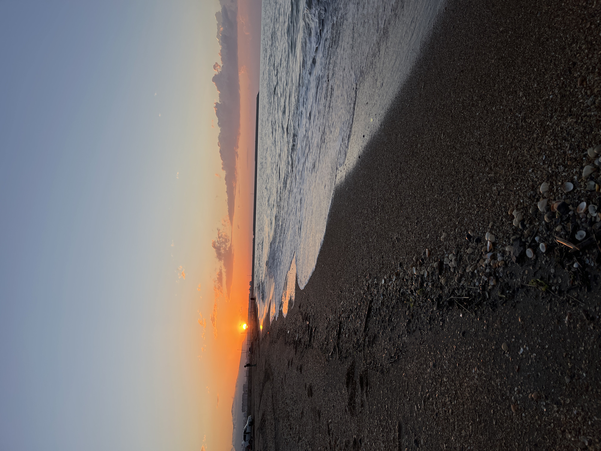 The image shows a sunset view on the beach in Kaspiysk, Russia. The sun is setting on the horizon, creating an orange glow on the sky and reflecting on the water. The beach is made of dark sand and pebbles, with some small shells scattered around. Waves are lapping onto the shore. There are a few figures of people on the beach near the sunset, and vehicles appear to be parked along the shore, possibly on a road. The sky is clear with some light clouds on the horizon.