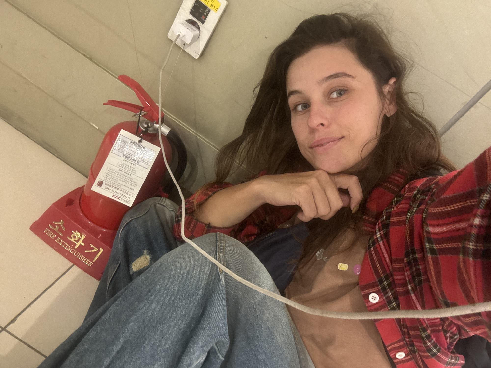 A young woman with long, dark, wavy hair is lying down and taking a selfie. She is wearing a red and black plaid shirt and distressed denim jeans. Her head is propped up on her hand, and she is looking towards the camera with a slight smile.
In the background, there is a red fire extinguisher with a label indicating it is a "FIRE EXTINGUISHER" in both Korean and English. Next to the fire extinguisher, a white electrical outlet with a charging cable plugged into it is visible on the wall. A yellow sticker with Korean text and "H1 #14" is also attached to the outlet. The floor appears to be tiled.
The scene suggests the woman is in an indoor public space, possibly an airport or a waiting area, given the presence of a fire extinguisher and the electrical outlet. The lighting appears to be artificial, and there are no strong indicators of the time of day or weather conditions. The woman's relaxed posture and smile convey a sense of ease or contentment. Visible text includes Korean and English labels on the fire extinguisher, information on a tag attached to it, and markings on the electrical outlet.