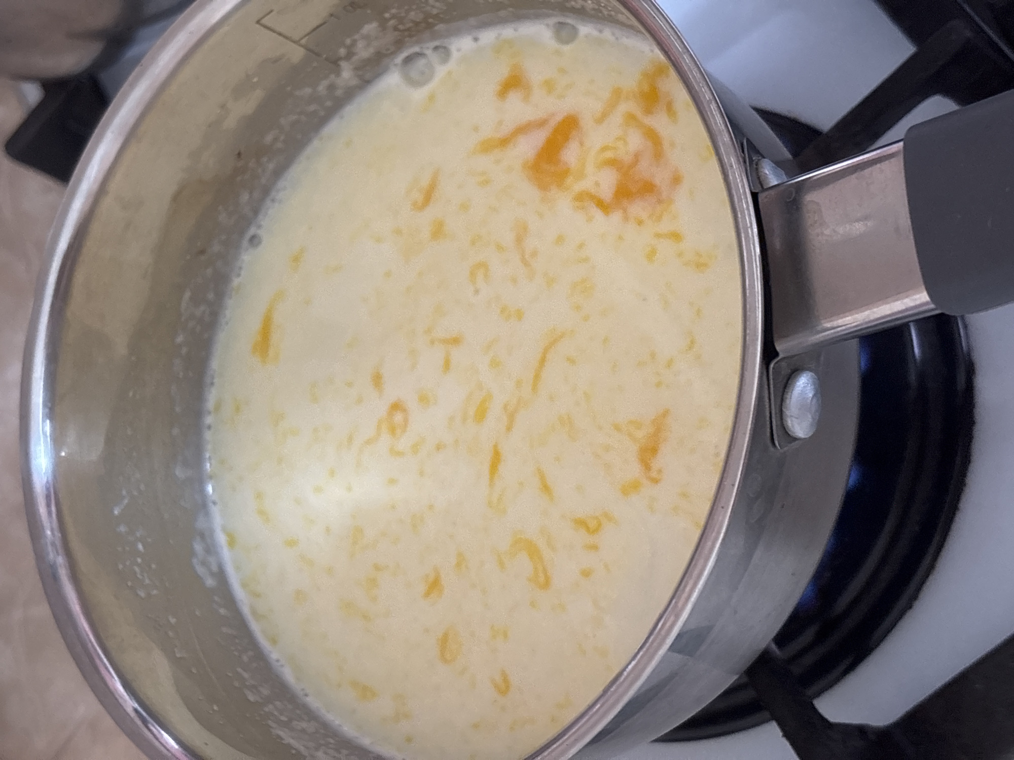 A close-up, overhead view of a stainless steel saucepan filled with a creamy, pale yellow liquid containing small, irregular pieces of bright orange, possibly citrus zest. The saucepan is placed on a gas stovetop, with a faint blue flame visible beneath the burner. The liquid appears to be gently simmering, with small bubbles on the surface. The handle of the saucepan, made of black plastic and metal, is visible in the foreground. The background is blurred but suggests a domestic kitchen environment with tiled countertops. There are no people or animals present. This scene is located in the city of Новый Кинер, Russia.
