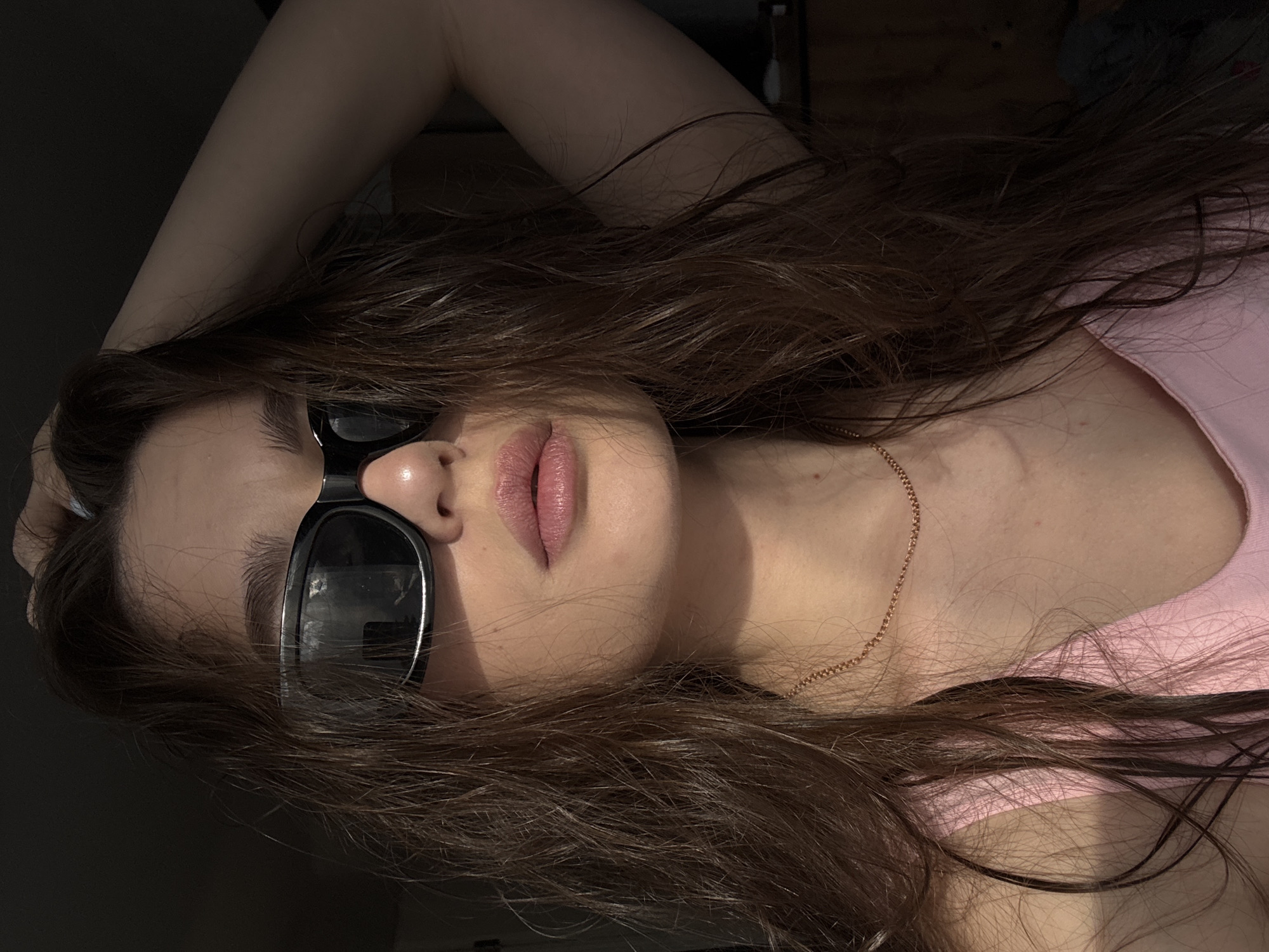 A young woman with long brown hair is lying down, wearing sunglasses and a pink shirt. A gold chain is visible around her neck. The location is not discernible from the provided image.