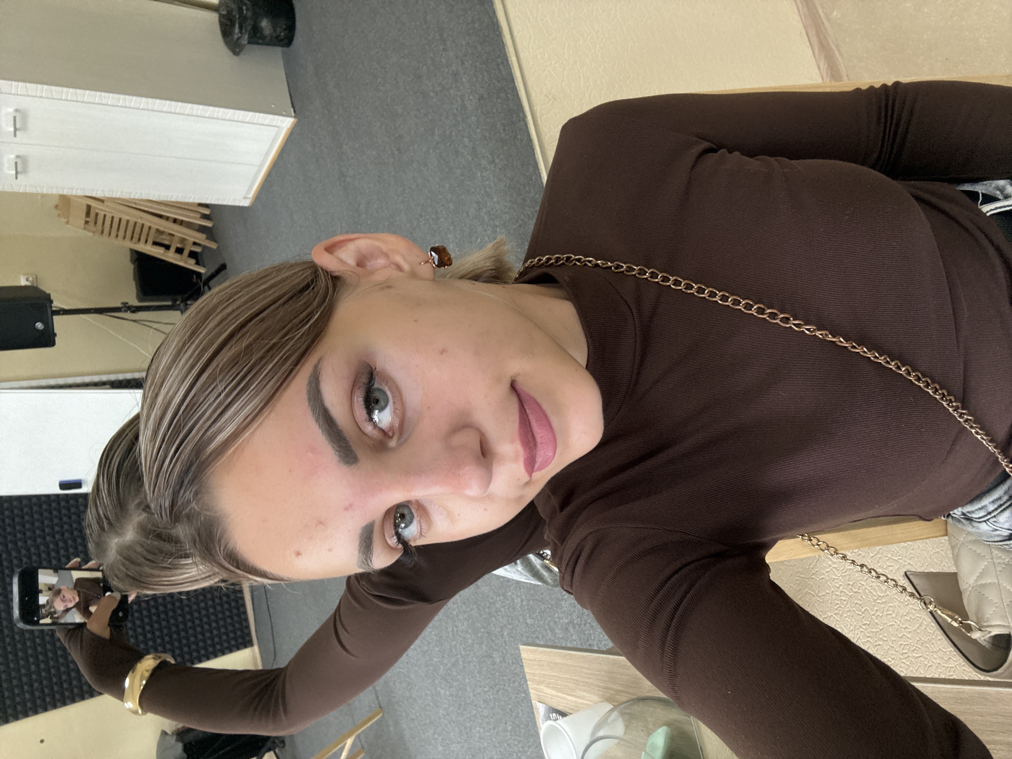 An indoor selfie is taken by a young woman with blonde hair, blue eyes, and makeup. She is wearing a brown turtleneck shirt and jeans. She is holding a smartphone in her left hand, which is visible in the frame as a reflection in the camera. A gold bracelet is on her left wrist. She is wearing brown earrings. A gold chain purse strap is draped over her shoulder. The background shows a studio-like setting with acoustic panels, a speaker on a stand, folded chairs, and a white cabinet. The floor is carpeted. The image does not provide any specific details that would allow for identification of the location within Yuzhno-Sakhalinsk, Russia.