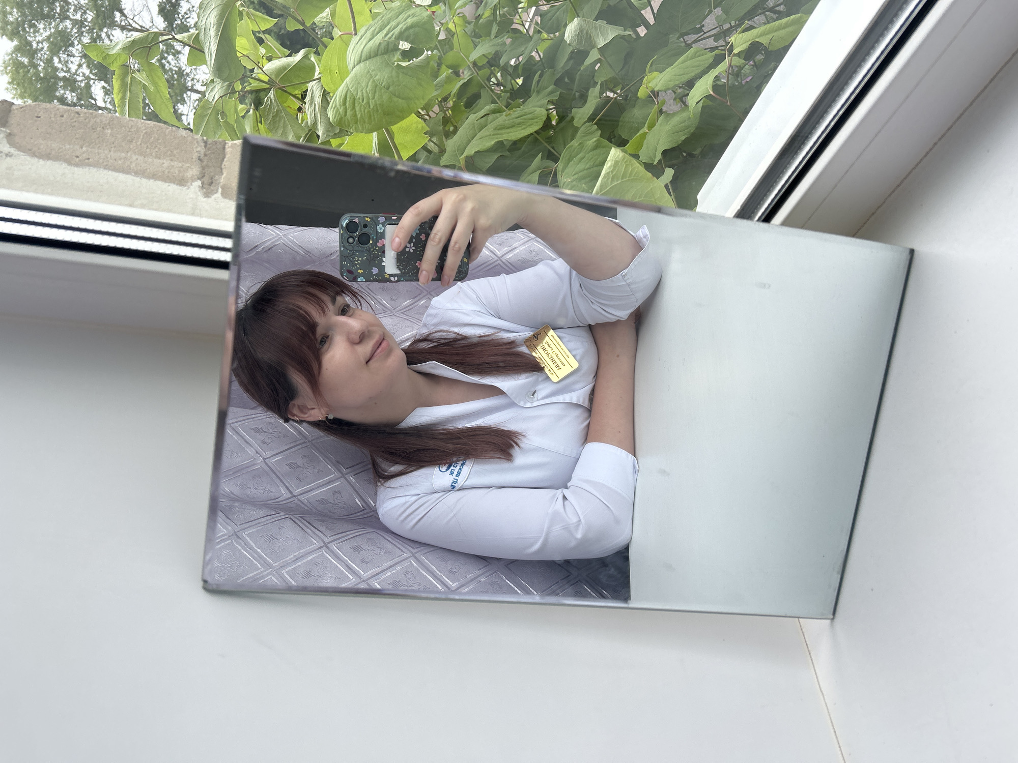 A mirrored image shows a young woman with shoulder-length, dark brown hair with bangs and pigtails. She is holding a smartphone with a floral case, seemingly taking a selfie. She is wearing a white collared shirt with a badge on her right breast; the badge appears to have text in Russian, and the text indicates the city of Фоки, Russia. The reflection shows the woman is lying on a quilted surface, while the mirror rests on a white surface. In the background, visible through a window, are green plants and a light-colored concrete structure. The photo appears to be taken indoors near a window.