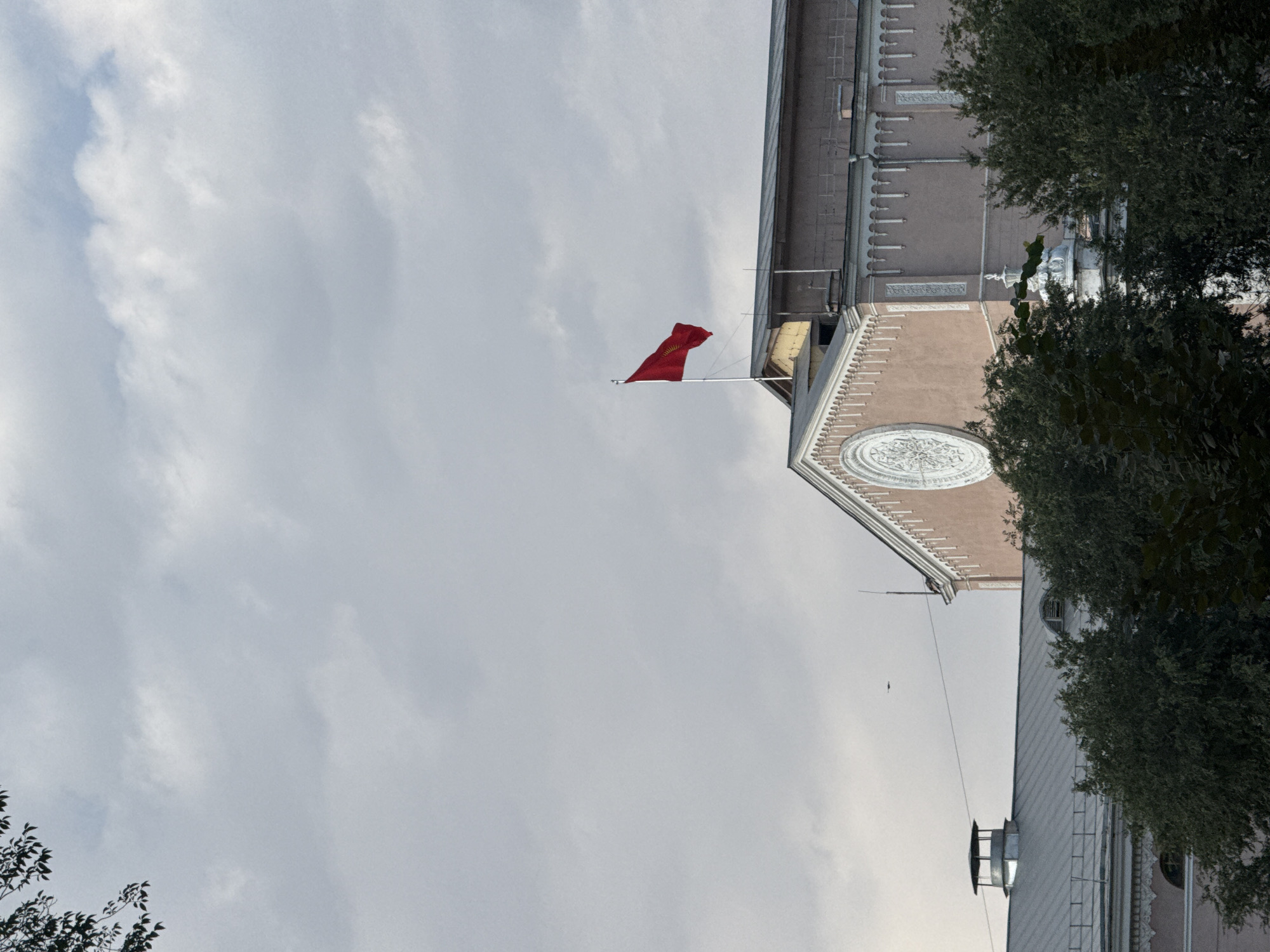 The image depicts a red flag with a yellow sun emblem on a flagpole atop a building. The building appears to be a large, ornate structure with a pink facade and decorative architectural elements, including a large circular ornament. Trees with dark green foliage are visible in the foreground, partially obscuring the lower portion of the building. The sky is overcast with gray clouds. In the distance, a small dark object, possibly an aircraft or drone, is visible against the sky. The scene is likely in Bishkek, Kyrgyzstan, given the prominent display of the Kyrgyz flag.