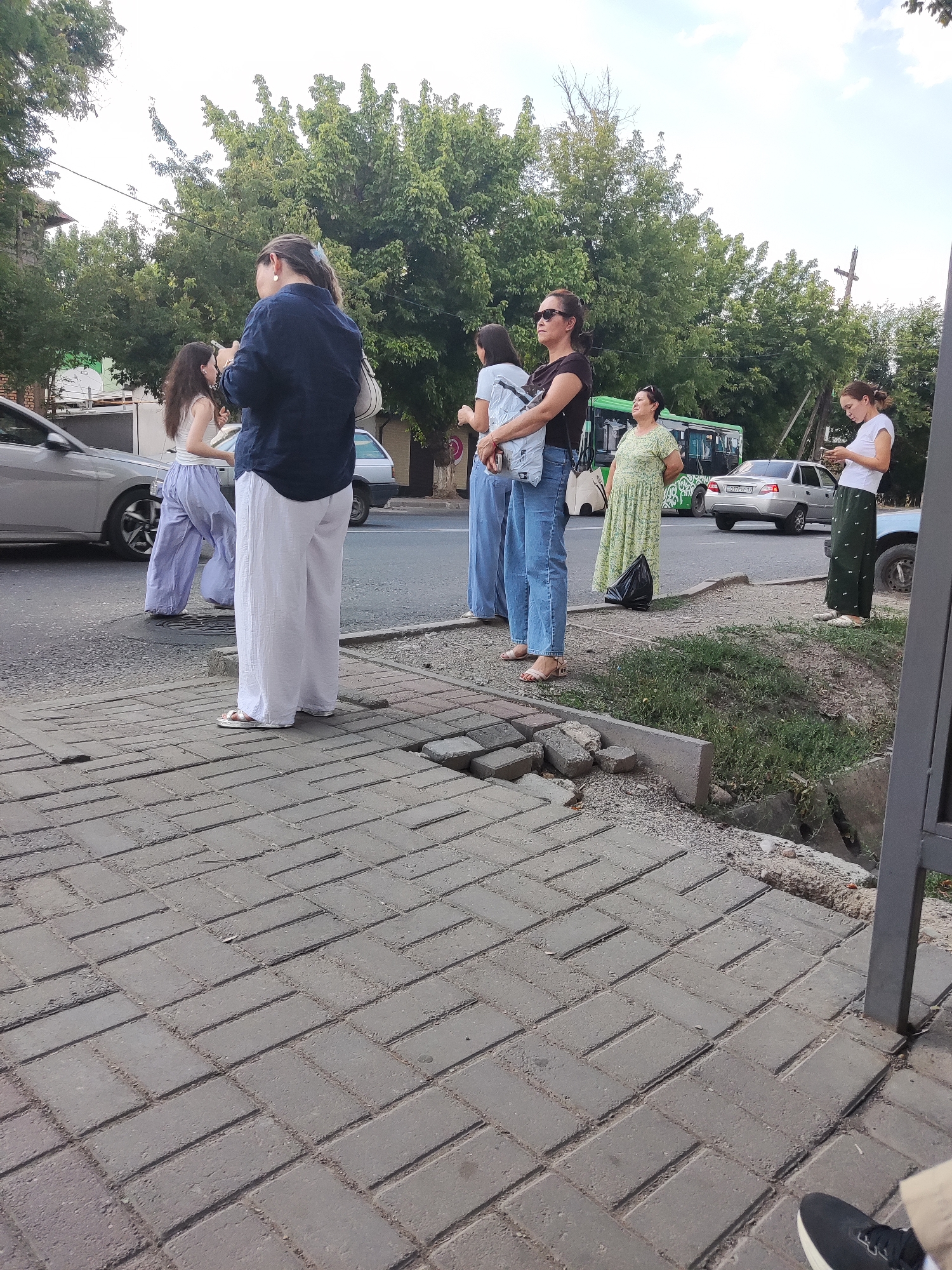 A group of people are standing on a sidewalk next to a street in Shymkent, Kazakhstan. The sidewalk is paved with gray rectangular bricks. In the background, there are trees, cars, and a bus. The sky is cloudy. One woman in the foreground is wearing a dark blue shirt and white pants, and she is holding a white purse. Another woman next to her is wearing a white tank top and light purple wide-leg pants. A third woman is wearing a black t-shirt and blue jeans. A fourth woman in a green dress is standing next to a black garbage bag. A fifth woman in a white t-shirt and green pants is looking at her phone. The scene appears to be a typical urban streetscape.