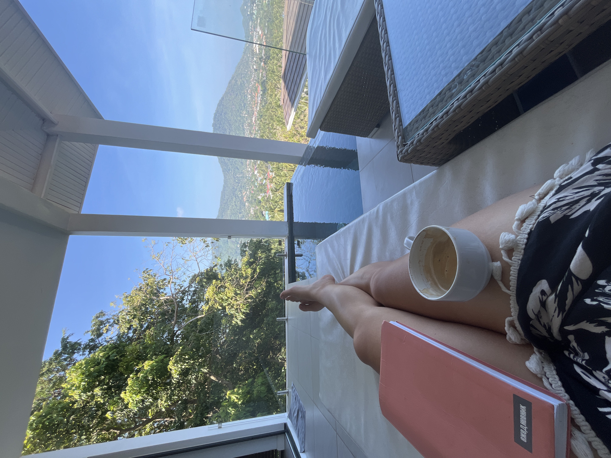 The image shows a person reclining outdoors on a lounge chair, possibly on a balcony. The person is holding a mug, presumably of coffee, and a book. The balcony overlooks lush green hills and trees. A swimming pool is visible in the foreground, and a table with chairs is nearby. The location is Baan Lamai, Thailand, suggested by the background scenery.