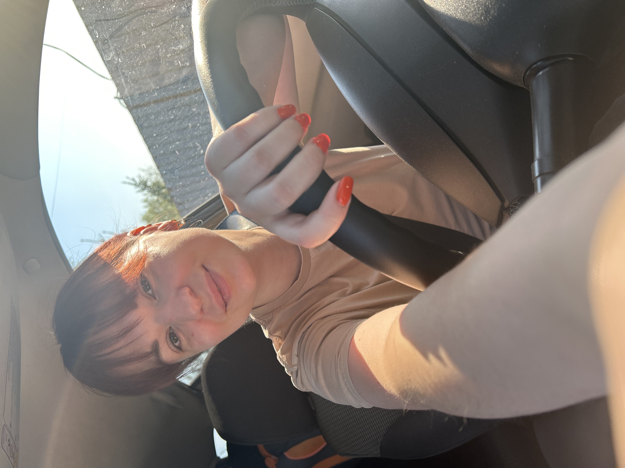 The image depicts a woman inside a car. She has brown hair with bangs and is wearing a beige t-shirt. Her fingernails are painted red. She is holding onto the steering wheel of the car. The background shows a view through the windshield, including a glimpse of a tree and sky. The location appears to be Фоки, Russia.