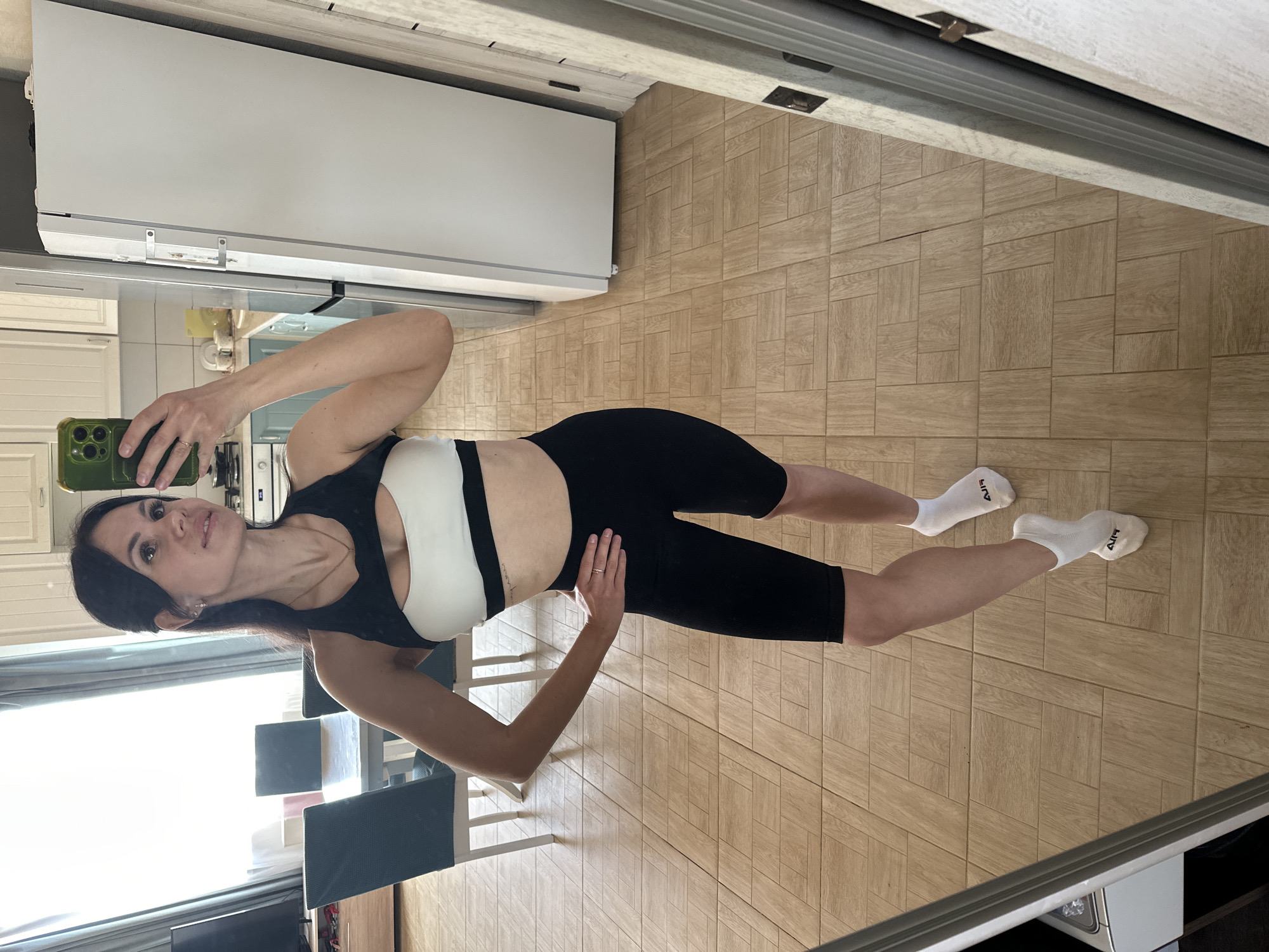 A woman is taking a selfie in what appears to be a kitchen. She is wearing a black and white sports bra and black athletic shorts, along with white socks. The kitchen has wooden floors and white cabinets. A dining table with gray chairs can be seen in the background. The location is Unknown, Unknown.