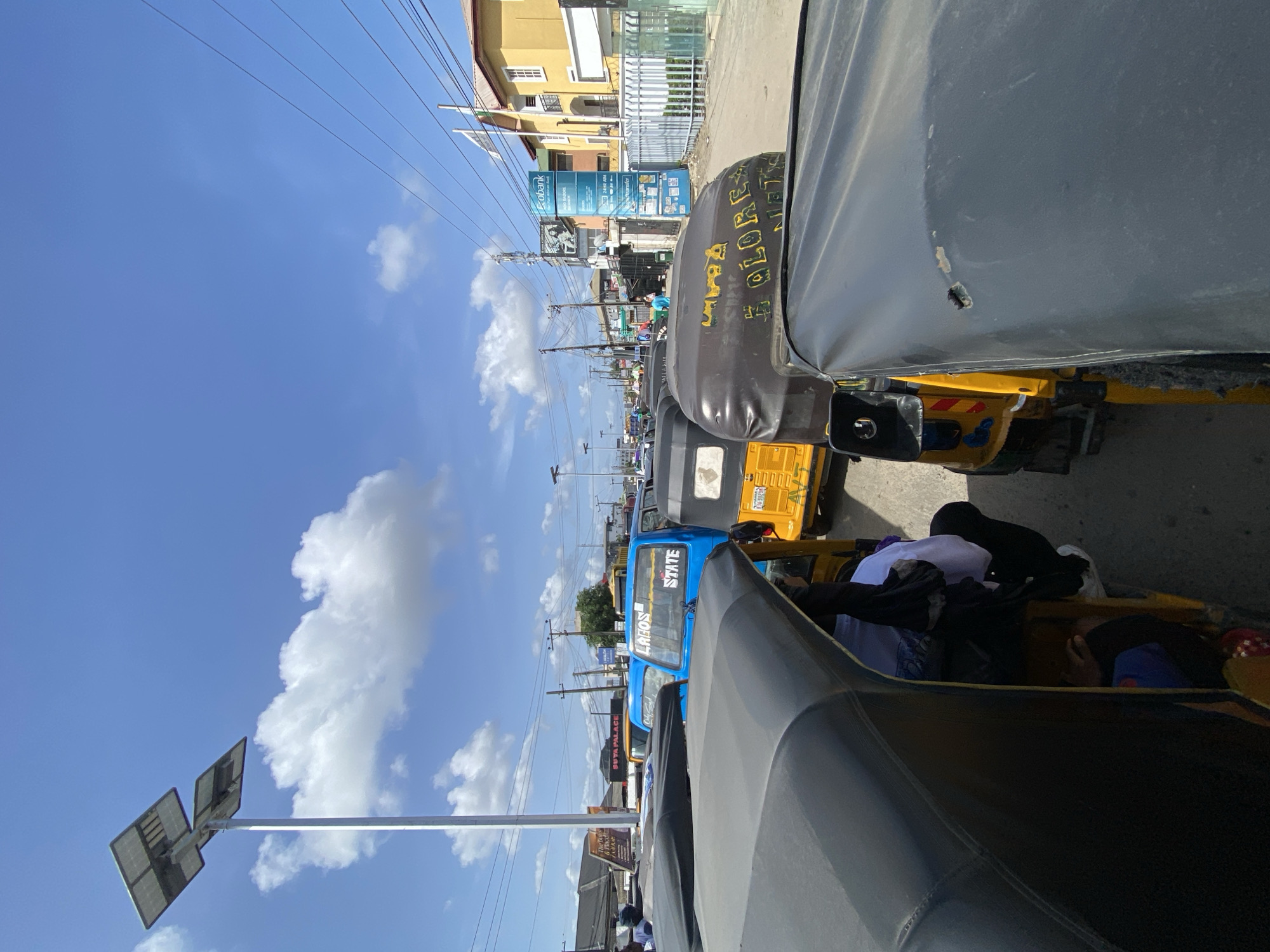 The image depicts a street in Ado, Nigeria, filled with parked or slow-moving "keke" or "tricycle" vehicles. The foreground is dominated by the covered canvas roof of a keke. Behind it are several other kekes in shades of blue, yellow and black, forming a line along the street. "LAGOS STATE" is clearly visible on the back of a blue keke. "LAGOS" is visible on the blue keke. Further down the street, buildings and utility poles line the road, with a cloudy blue sky visible above. The scene appears to be a typical urban street in Nigeria, possibly during a busy time of day. Some passengers appear to be inside the kekes. There is writing on the roof of one of the kekes, including part of the word "GLORY."