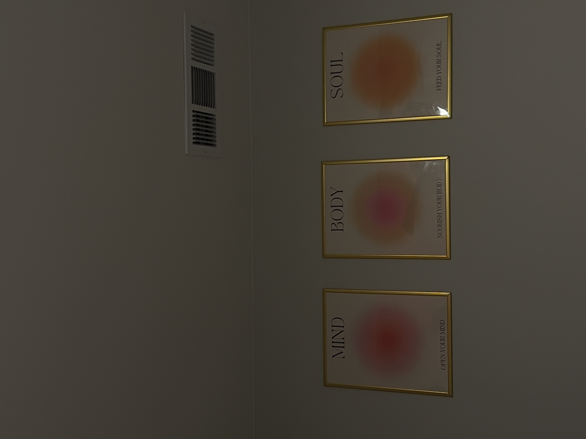 This image captures a dimly lit interior wall, likely in Austin, United States of America, featuring both functional and decorative elements. To the left, a white rectangular HVAC vent is visible, integrated into the wall, indicating an indoor climate-controlled environment.
On the right side of the frame, three identical framed prints are stacked vertically. Each print is encased in a golden frame and features a blurred, gradient circle resembling an aura, along with text.
The topmost print has an orange and yellow gradient circle. Vertically aligned on the left side of the print is the word "SOUL," and on the right, the phrase "FEED YOUR SOUL" is written, also vertically.
The middle print displays a pink and magenta gradient circle. On its left, the word "BODY" is vertically positioned, while the right side reads "NOURISH YOUR BODY" vertically.
The bottom print features a red and pink gradient circle. Vertically on the left, the word "MIND" is visible, and on the right, the phrase "OPEN YOUR MIND" is written vertically.
The overall impression is one of mindfulness and self-care, emphasizing the importance of nurturing different aspects of oneself. The low lighting suggests either evening hours or a room with subdued illumination. No specific activity is depicted, making the scene a static display.