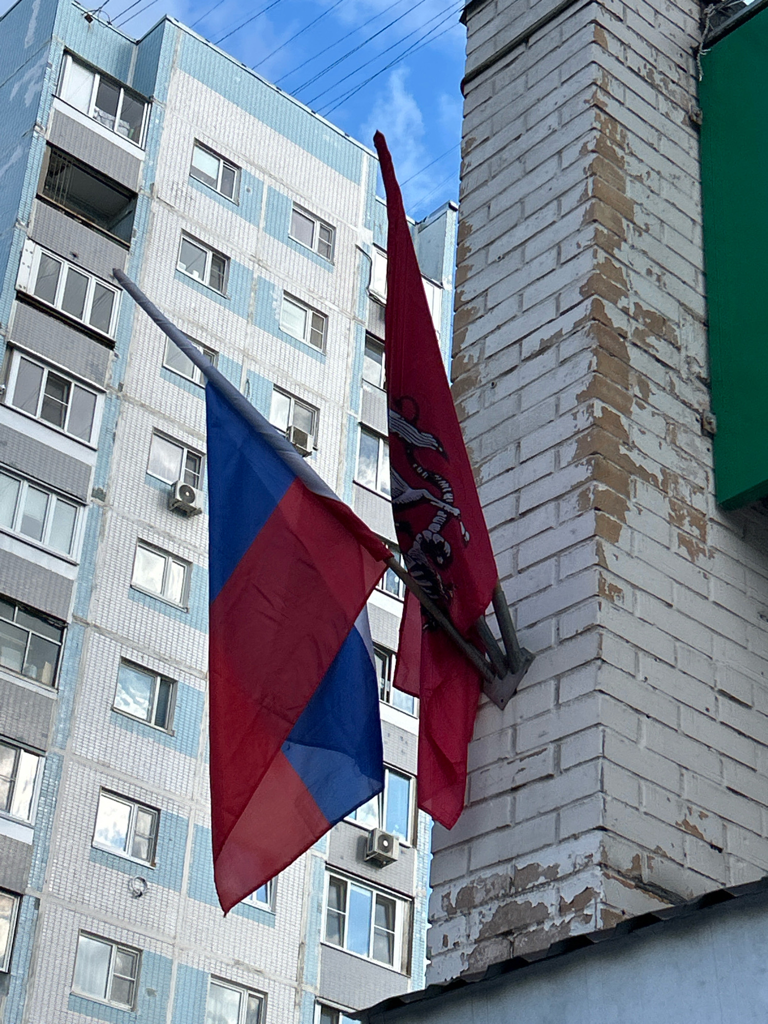 In Moscow, Russia, a close-up, eye-level shot captures two flags displayed on the exterior of a weathered brick building. The Russian tricolor flag, with its distinct white, blue, and red horizontal stripes, hangs to the left, partially obscuring a light blue and white tiled apartment building in the background. To the right, a red flag features a prominent black emblem, possibly a coat of arms or insignia. The brick building itself shows signs of age, with peeling white paint revealing the underlying brickwork. The flags are attached to metal poles that are mounted to the corner of the brick structure. The sky above is a mix of blue and clouds.
