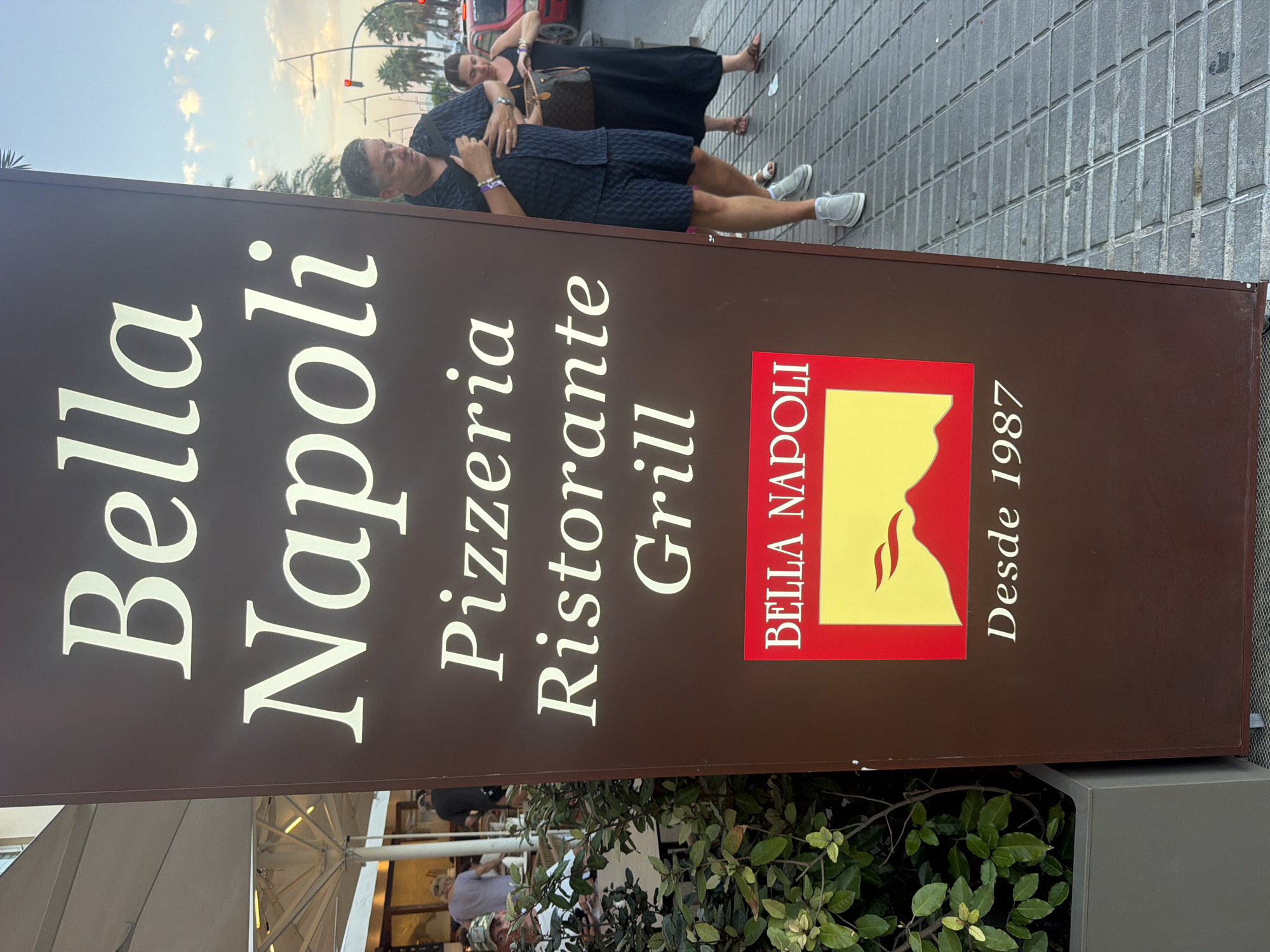 A vertical brown sign for "Bella Napoli Pizzeria Ristorante Grill" stands on a sidewalk in Ibiza, Spain. The sign is illuminated with white lettering. At the bottom of the sign is a red and yellow logo with the text "BELLA NAPOLI" and an image of a volcano erupting. Below the logo, the year "Desde 1987" is displayed. To the right of the sign, a man in a dark patterned shirt and shorts, wearing grey slip-on shoes, is adjusting his shirt. A woman in a black dress and sandals, carrying a brown handbag, stands behind him. In the background, palm trees, a street light with a red signal, and a red car are visible. To the left of the sign, people are seated at tables under a large white umbrella, with a potted plant in the foreground.