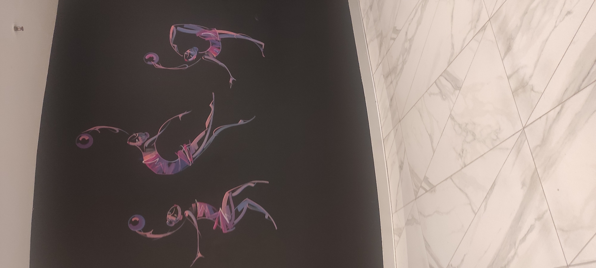 The image shows a wall decorated with stylized art of three rhythmic gymnasts in motion. Each gymnast is depicted with vibrant, multi-colored lines creating a sense of fluidity and dynamism as they perform. The gymnasts are holding and manipulating spherical objects. The background of the artwork is a dark, solid color. Below the wall, a floor with a marble-like pattern is visible, laid out in large, rectangular tiles. The lighting in the image suggests an indoor setting. There is no indication in the image that this scene is located in Moscow, Russia.