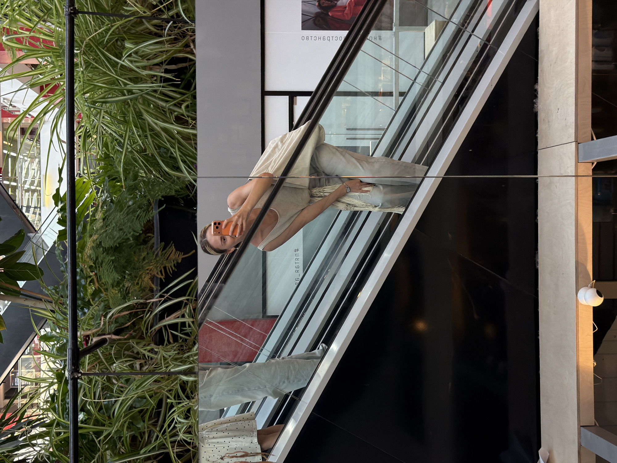 This image captures a reflection within a modern indoor commercial space, likely a shopping mall, located in Khimki, Russia. The primary subject is a woman, reflected as if standing on an upward-moving escalator, taking a mirror selfie with an orange-cased smartphone held in her right hand. She is wearing a light-colored sleeveless top, light blue/grey wide-leg pants, and a light-colored fringed scarf or shawl draped around her waist. A watch is visible on her left wrist.
To her left in the reflection, two other individuals are partially visible on the escalator: one person's lower legs and feet wearing a light-colored, polka-dotted dress or skirt, and another person's lower legs in light-colored pants.
The reflected environment above the escalator features multiple planters filled with lush green foliage, including variegated plants resembling spider plants, and other dense green bushes. A black metal railing runs horizontally above these plants. Beyond the escalator to the right, white and grey wall panels are visible, with some mirrored Cyrillic text ("ООС1О9НСLВО") and a hashtag ("#НО") on a white surface, along with a partial image of a figure in red clothing.
In the bottom foreground of the actual photograph, a light wooden beam or ledge is visible, with a white dome-shaped surveillance camera mounted on it, facing towards the lower right. Below this beam, a dark, reflective surface is present.