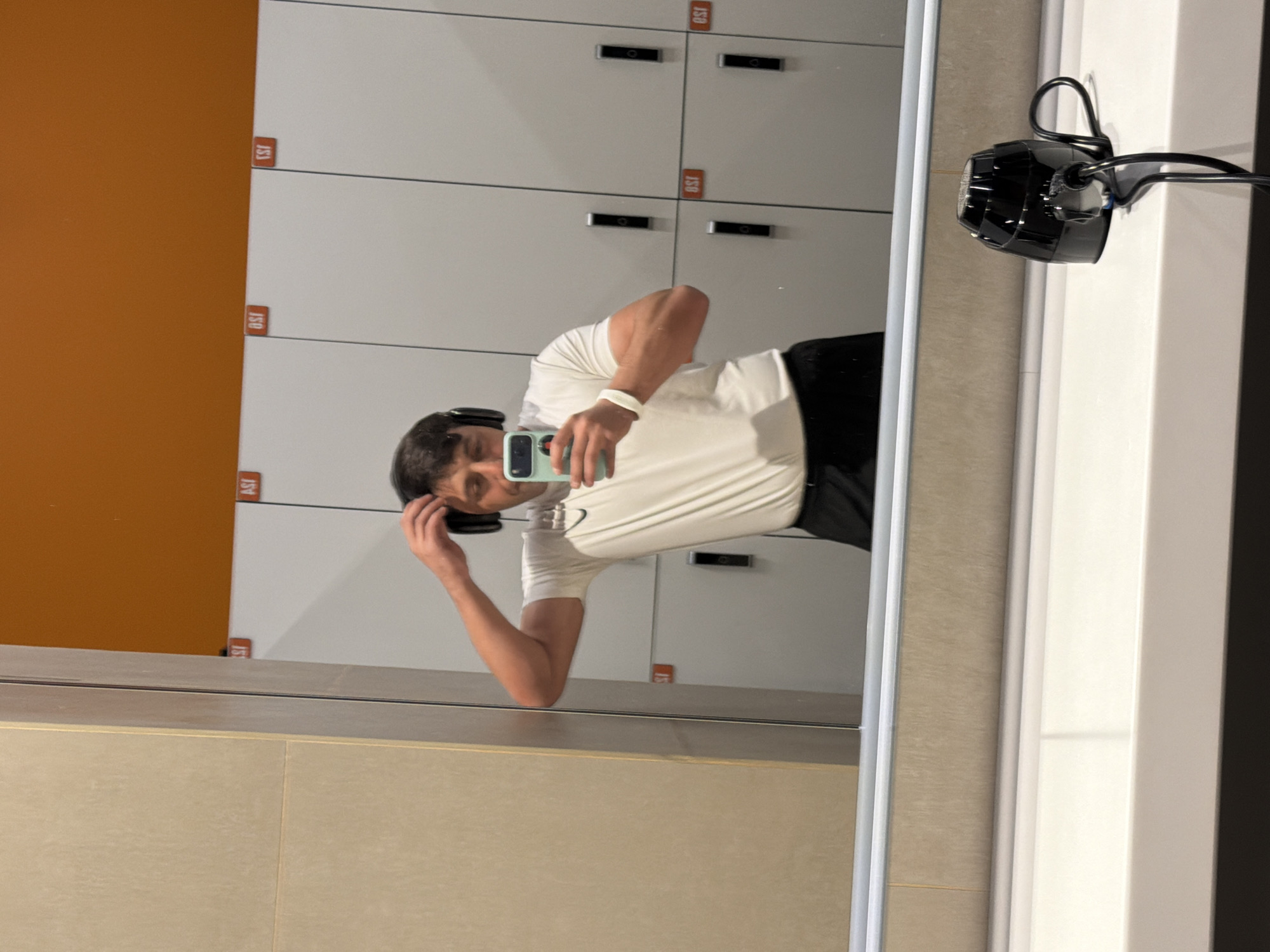 A rotated (90 degrees clockwise) mirror selfie captures a young man in what appears to be a locker room or changing area, possibly within a gym in Krasnoyarsk, Russia. The man is reflected in the mirror, wearing a white short-sleeved t-shirt, black shorts or pants, and black over-ear headphones resting around his neck. A white wristband is visible on his left wrist, and he holds a light blue smartphone to take the photograph. His expression is neutral as he looks towards the phone. His right arm is bent at the elbow, with his hand near his head, possibly adjusting his hair.
The background, reflected in the mirror, features white lockers with black handles or locking mechanisms. Visible red-orange labels with black text are affixed to the locker doors, displaying numbers such as "J53", "J50", and "J51". An orange wall is visible to the left of the lockers' reflection. The lower portion of the image shows a reflection of a beige or light brown tiled floor. On the right side of the mirror, a black, multi-faceted object, possibly a light fixture, speaker, or camera, with visible wires, is mounted to the wall. The scene is brightly lit, indicative of an indoor facility.