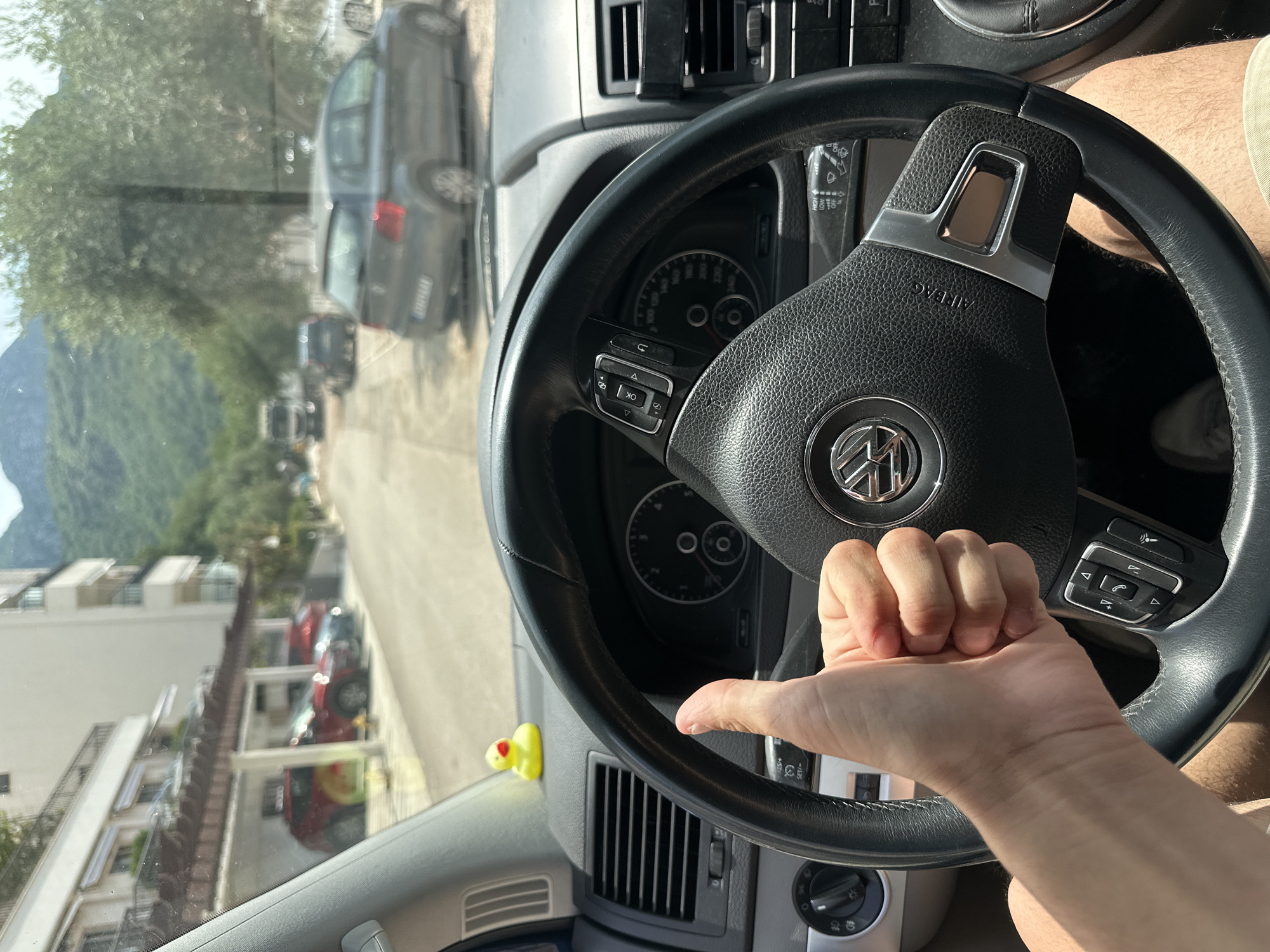 The image, rotated 90 degrees clockwise, presents a first-person view from the driver's seat of a car, looking out the windshield onto a sunny street scene.
In the foreground, the car's interior is visible, dominated by a black leather steering wheel featuring a prominent Volkswagen (VW) logo in the center and "AIRBAG" inscribed below it. The steering wheel is equipped with various control buttons on both sides, including "OK," directional arrows, phone icons, and volume controls on the left, and cruise control settings on the right. Behind the steering wheel, parts of the instrument cluster are discernible, showing a speedometer marked up to 240 km/h, and a tachometer. Dashboard air vents are visible on the dashboard, along with the light control knob and a wiper stalk displaying text like "HIGH," "LOW," and "OFF." A small, bright yellow rubber duck toy with a red beak is perched on the dashboard near the windshield.
A person's right hand, making a "thumbs-up" gesture, rests casually on the steering wheel. Part of a person's leg, likely the driver's, is visible on the far right side of the frame.
Through the windshield, a street scene unfolds under bright daylight and clear skies. Several cars are parked along the road, including a dark-colored sedan or hatchback positioned directly in front of the viewer's car. Buildings, appearing to be multi-story apartments or houses, line both sides of the street. Lush green trees and other foliage are scattered along the road and between the structures. In the distant background, verdant mountains rise prominently, indicating a mountainous landscape.
The bright lighting suggests it is midday or early afternoon. The overall setting is a picturesque, possibly residential or tourist area, consistent with the specified location of Trap, Montenegro. The thumbs-up gesture might convey a positive sentiment or satisfaction with the moment or location.