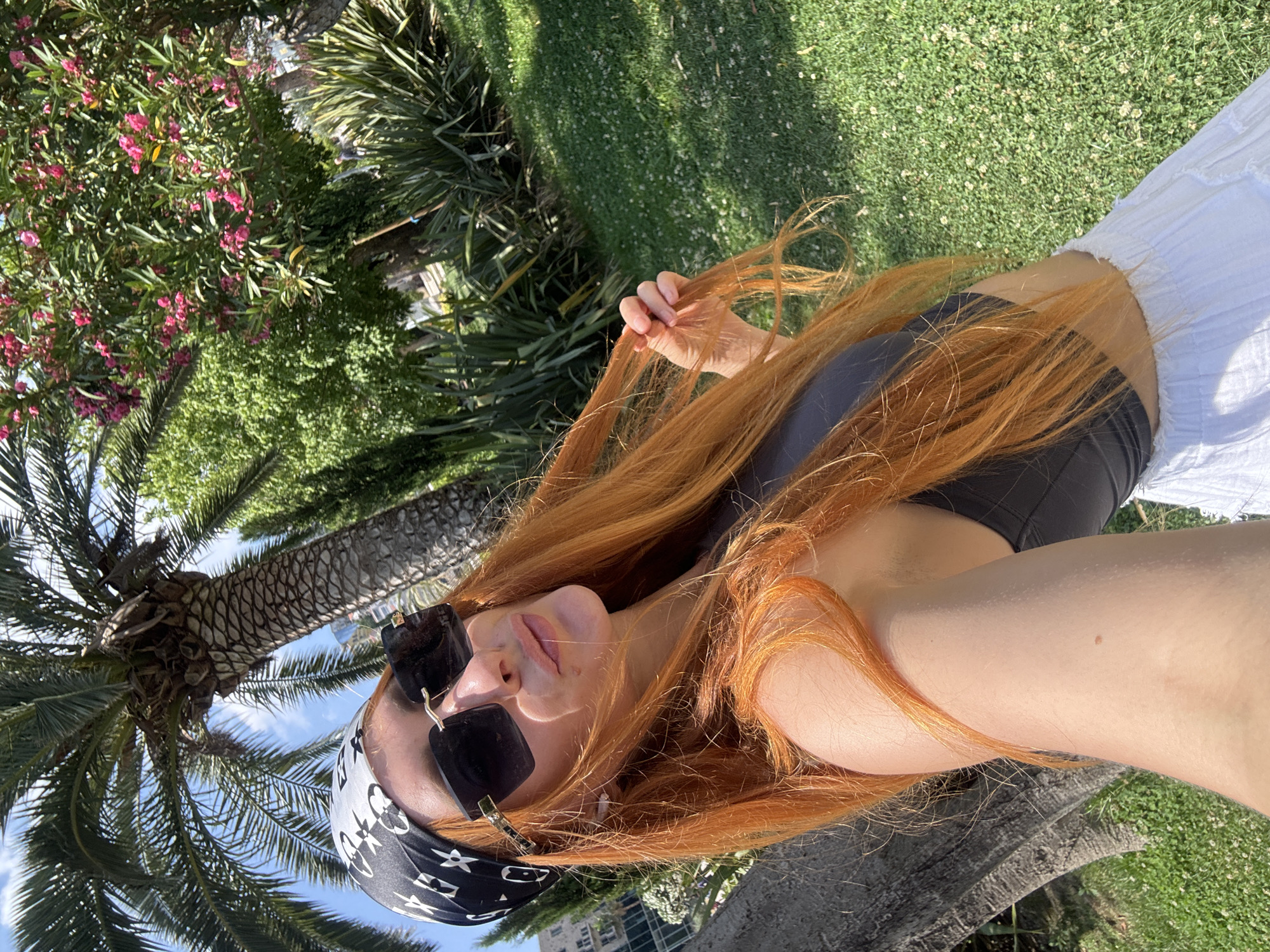 A woman with vibrant orange hair, wearing a black bandana with a white pattern and dark sunglasses, takes a selfie outdoors. She is dressed in a black top and a white skirt. Her right hand is holding a strand of her hair. The background features a palm tree, lush green grass with small white flowers, and foliage. A building is partially visible in the distance. The overall setting appears to be a park or garden in a sunny, urban environment, possibly Budva, Montenegro, given the presence of Mediterranean flora.