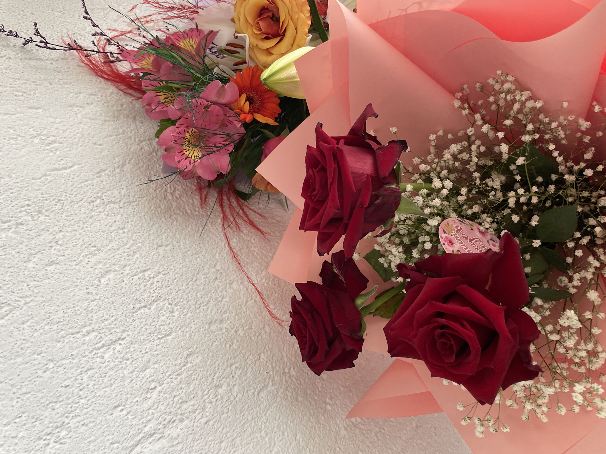 A bouquet of flowers rests against a textured, off-white surface. The flowers include red roses, small white flowers, and other flowers in shades of pink, orange, and yellow. The bouquet is wrapped in pink paper. The background appears to be a wall with a rough texture. There is no indication of the location being Zlatograd, Bulgaria or any presence of people, actions, or interactions in the provided image.