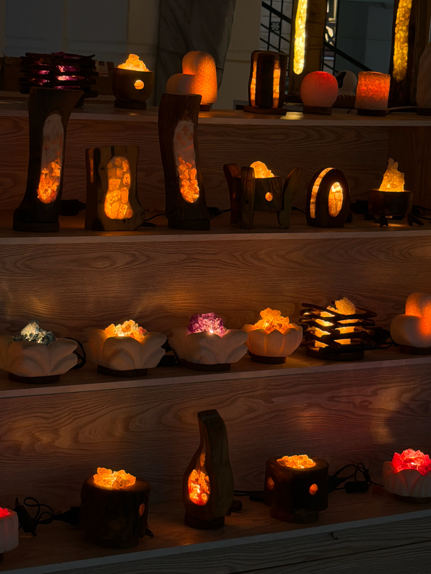 The image depicts a dimly lit retail display showcasing an assortment of decorative salt lamps. The lamps are primarily made of natural wood and feature carved openings through which a warm, orange glow emanates from the salt crystals within. The display is organized on wooden shelves, suggesting an indoor environment, possibly a shop or market stall. Various shapes and sizes of lamps are visible, including cylindrical, irregularly shaped, and bowl-like designs, some with a lotus flower motif. In the foreground, several smaller lamps are arranged, while larger, more prominent pieces occupy the upper shelves. The overall atmosphere is warm and inviting, accentuated by the soft illumination. The scene is devoid of any human presence. The reference to Sharm El Sheikh, Egypt, implies this retail setting is located within that city.